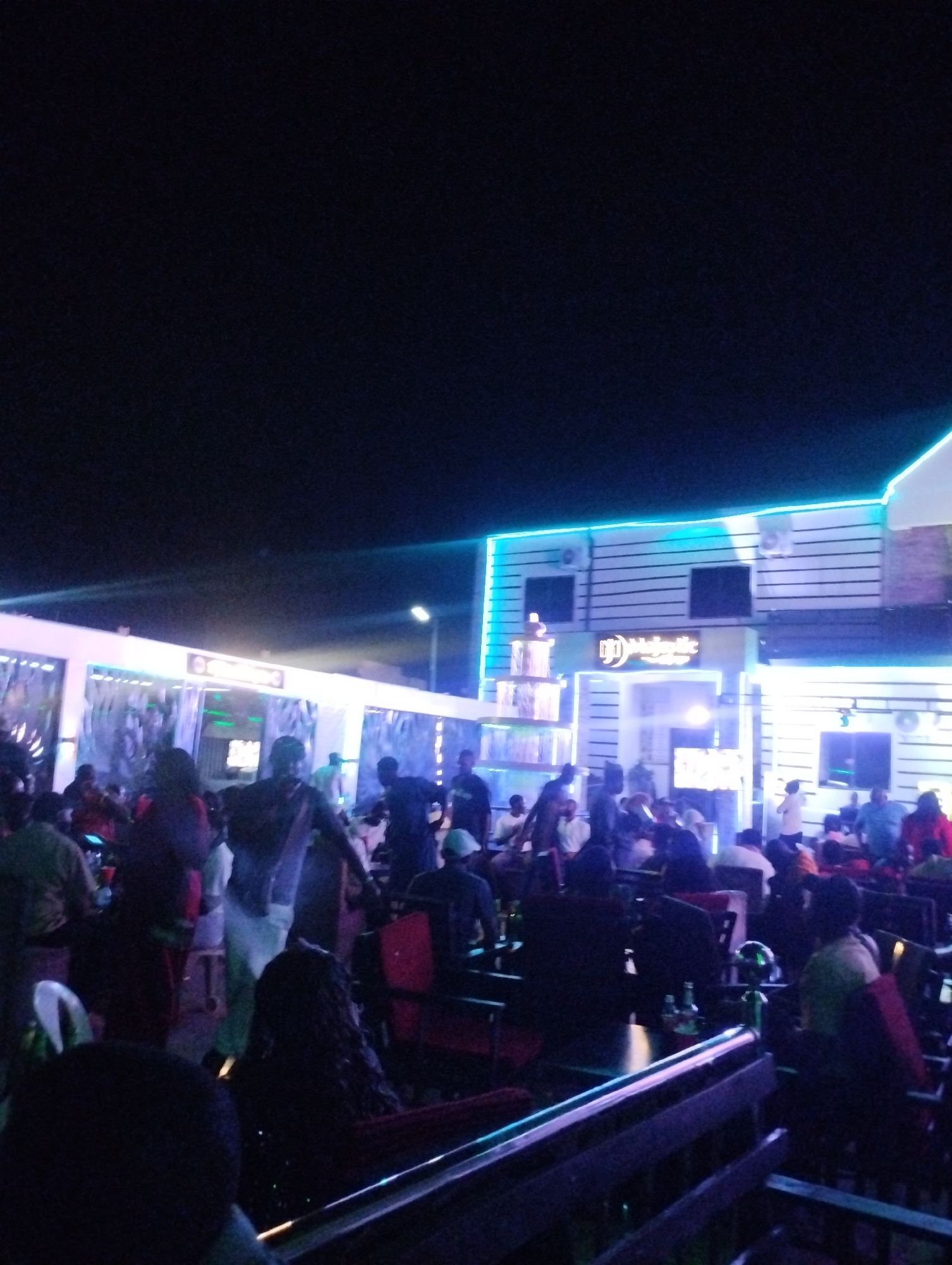 The image depicts an outdoor nighttime scene in Tumfure, Nigeria. A crowded outdoor bar area is filled with people seated at tables and mingling. The bar is illuminated with blue neon lights and features a sign that reads "Majestic." Several individuals are standing and walking around, while others are seated at tables with drinks. The environment appears to be a lively social setting. The sky is dark.