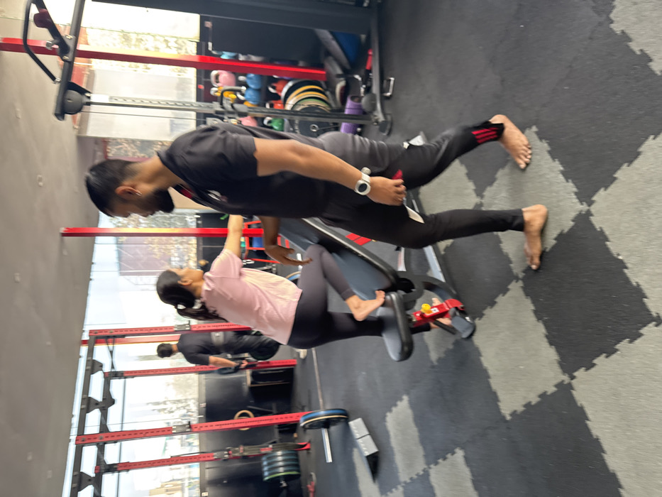 A man and a woman are in a gym. The woman is lying on her back on a workout machine with her feet in the foot holds. The man is standing next to her. He is wearing a black t-shirt and black pants. The woman is wearing a pink t-shirt and dark leggings. There are other people in the background. The gym has weights, machines, and other equipment. The floor is gray and black.