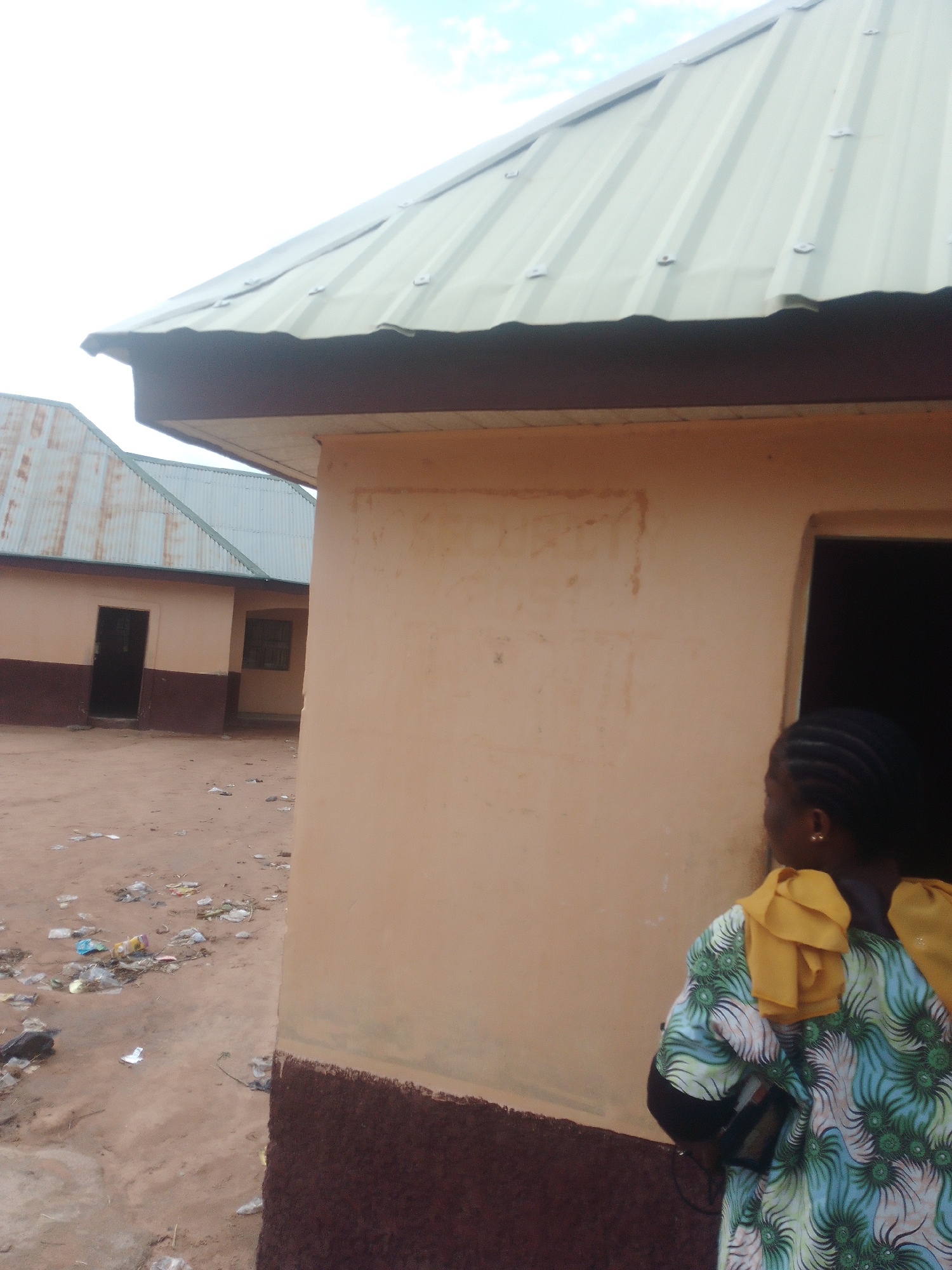 A medium shot captures a woman standing with her back to the camera, facing a light-colored building with a dark brown base. She wears a colorful, patterned garment with yellow accents. Her hair is styled in braids, and she has an earring in her visible ear. The building has a tan-colored wall and a dark brown foundation. To the left of the woman and the building, another similar building is visible with a corrugated metal roof and a dark-colored door. The ground between the buildings is unpaved and littered with debris. The sky above is partly cloudy. The scene appears to be outdoors during the daytime.