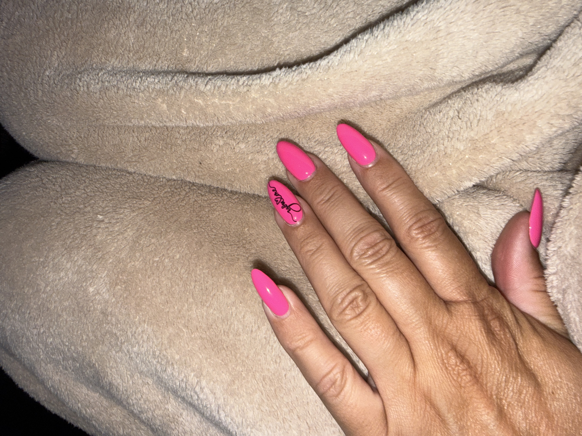 The image shows a person's hand with bright pink, almond-shaped acrylic nails resting on a soft, beige, fuzzy blanket. The hand is positioned in the lower right quadrant of the frame. The lighting suggests an indoor setting with soft, ambient light, possibly in the evening or a dimly lit room. The focus is primarily on the nails and the texture of the blanket.
Notable details include a black cursive script design on the ring finger nail, which appears to spell out "RubyLane". The skin on the hand shows some texture and is tanned. The blanket is folded, creating soft folds and shadows, contributing to the cozy and intimate feel of the image. No specific time of day or weather cues are discernible, and the location is given as Fairview Township, United States of America. The overall impression is one of personal care and attention to detail, with the vibrant nails standing out against the neutral background.