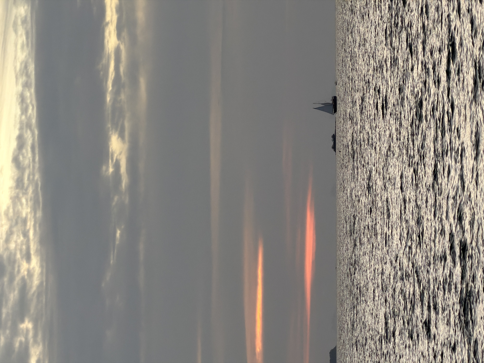 The image depicts a tranquil scene of the Gulf of Thailand, likely near the coast of Ko Samui District, Thailand, during twilight or sunrise. The sky is overcast with gray clouds, but a streak of warm, orange-pink light pierces through the clouds near the horizon, suggesting the sun is either setting or rising. The sea is moderately choppy, reflecting the muted colors of the sky.
In the middle ground, a sailboat with two white sails is prominently featured, sailing to the right of the frame. To the left of the sailboat, a larger, dark vessel, possibly a freighter or a larger boat, is visible. Further to the left, a distant landmass or island can be faintly seen on the horizon. There are no people clearly visible in the image. The overall atmosphere is serene and slightly melancholic due to the subdued lighting and cloudy conditions.