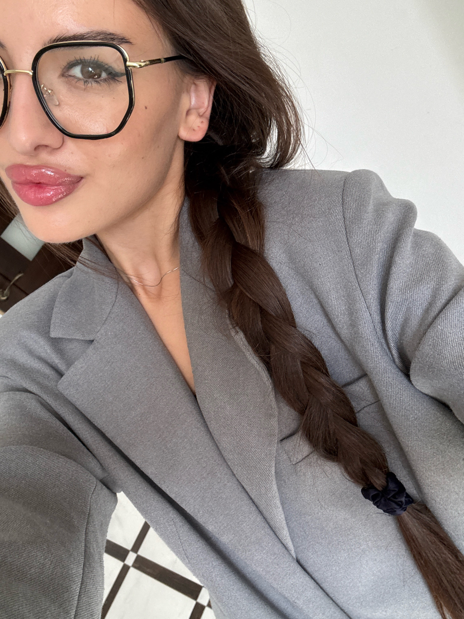 The image shows a close-up of a woman's face and upper body. She has dark brown hair in a braid, is wearing large, dark-rimmed glasses, a grey blazer, and has glossy pink lipstick on. The background is a plain white wall, with a hint of a checkered floor pattern visible at the bottom. There is no indication of the location being Chișinău, Moldova.
