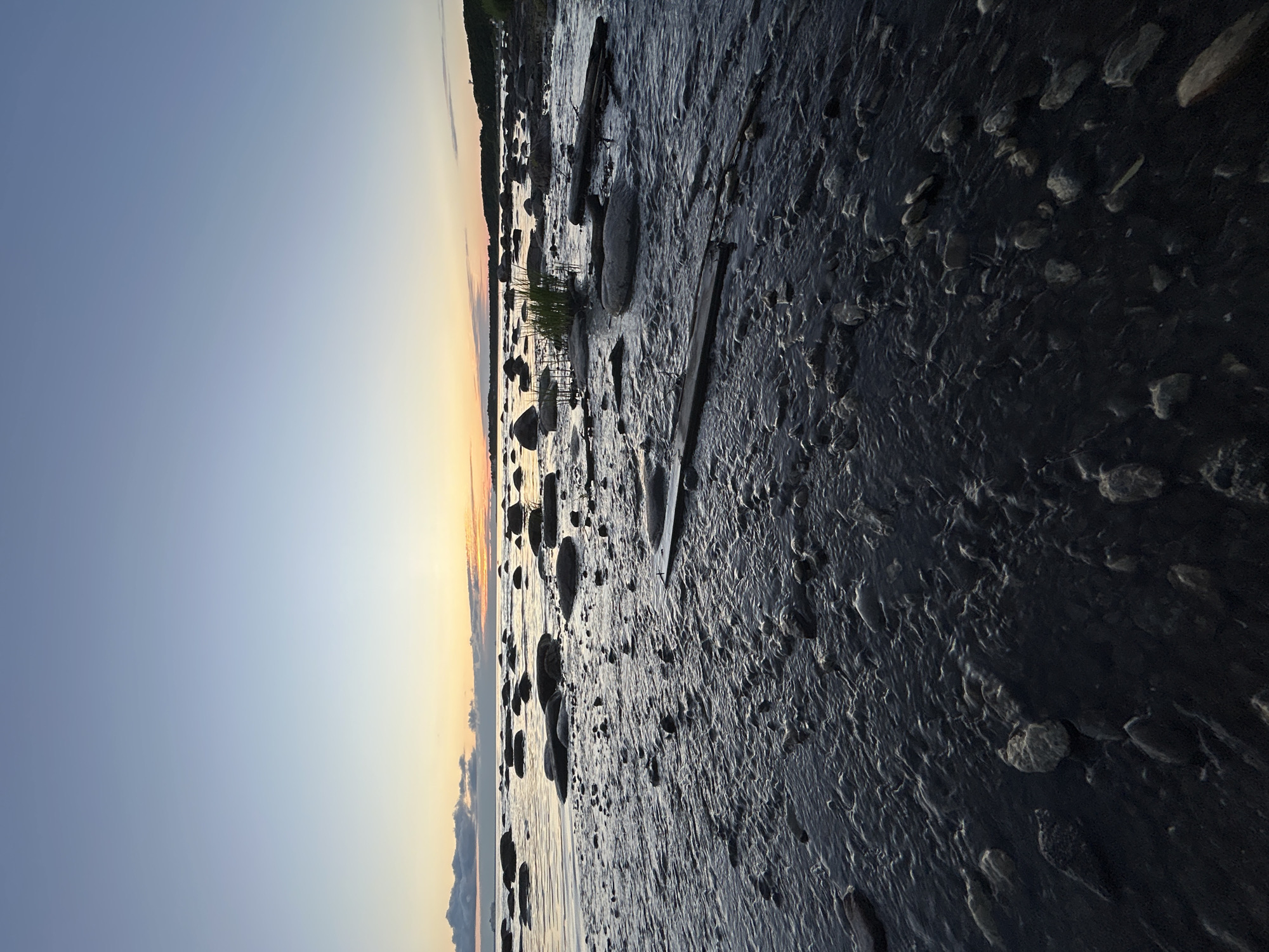 This is a vertical, eye-level shot of a rocky shoreline at dusk in Haabneeme alevik, Estonia. The foreground is dominated by dark, wet rocks and pebbles interspersed with shallow, rippling water that reflects the muted tones of the sky. In the middle ground, larger, smoother rocks emerge from the water, with some sparse grass growing between them. A few pieces of driftwood are scattered among the rocks. The horizon features a calm body of water, likely the sea, with a line of dark land and trees visible in the distance. The sky above transitions from a pale blue at the top to warmer hues of orange and yellow near the horizon, indicating the setting sun. There are a few scattered clouds, some dark and low-lying on the left, and lighter wisps of cloud further out. No people or animals are present. The overall atmosphere is serene and natural.