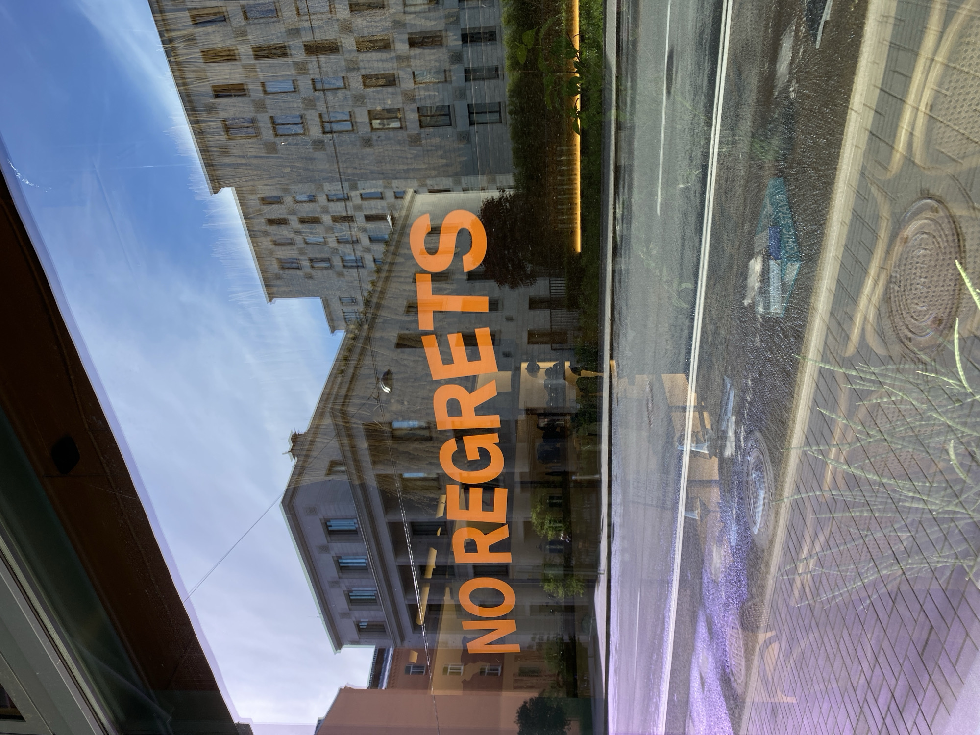 The image depicts a street scene in Saint Petersburg, Russia, reflected in a glass window with the orange words "NO REGRETS" overlaid. The reflection shows a cityscape with several multi-story buildings of varying heights and architectural styles. A road with painted white lines runs parallel to the buildings, and a sidewalk with patterned paving stones is visible on the far right. A manhole cover is embedded in the sidewalk. The sky above is clear and blue. The window frame is visible on the left side of the image.
