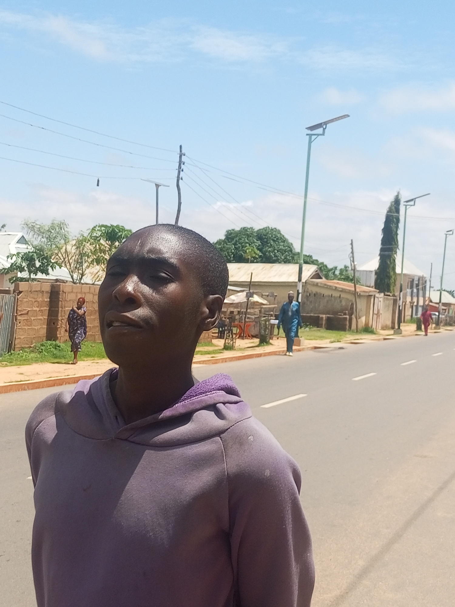 The image depicts a close-up of a young person's face and upper body, wearing a purple hooded sweatshirt. They are looking upwards and to the left of the camera, with their mouth slightly open. The background is blurred but shows a street scene. In the middle ground, a paved road runs horizontally, with white lane markings. To the left of the road, there is a brick wall and some greenery, with a woman in a patterned dress walking away from the camera. Further back, buildings and trees are visible. To the right of the road, a man in a blue traditional outfit is walking towards the camera. Several solar-powered streetlights are visible along the road. The sky is blue with scattered clouds. The image appears to have been taken outdoors during the daytime. The scene is consistent with a street in Tempure, Nigeria, characterized by its infrastructure, architecture, and vegetation.