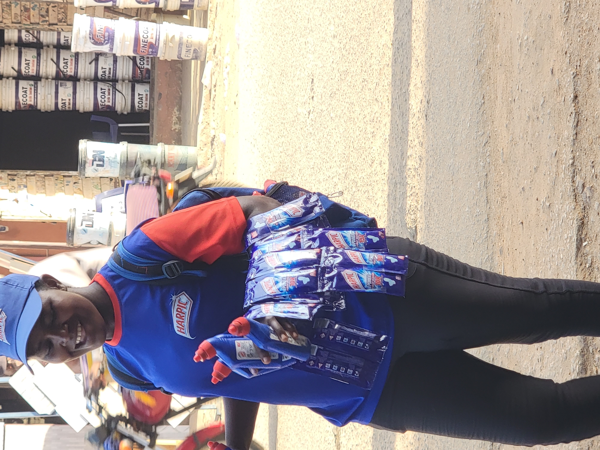 A person, likely a woman, is depicted standing outdoors, smiling warmly towards the camera. She wears a blue cap and a blue t-shirt with red accents, both prominently displaying the "HARPIC" logo, paired with black trousers. Attached to her front, likely via a harness or modified backpack, she carries numerous Harpic brand cleaning products, including multiple small blue sachets labeled "HARPIC POWER TOX" and several larger blue bottles with red caps. This setup suggests she is a street vendor or a brand promoter for Harpic products.
The scene is set on a sunny day, with bright daylight illuminating the area, implying clear weather. In the background, stacks of white plastic buckets are visible, many branded with text such as "FINECOAT," "ECOAT," "NECOAT," and "Acrylic Emulsion," indicating the presence of a paint or hardware store. The ground beneath her is a light-colored, paved surface, typical of a street or market in an urban or semi-urban environment like Maiburiji, Nigeria. Her pleasant smile conveys a friendly and approachable demeanor.