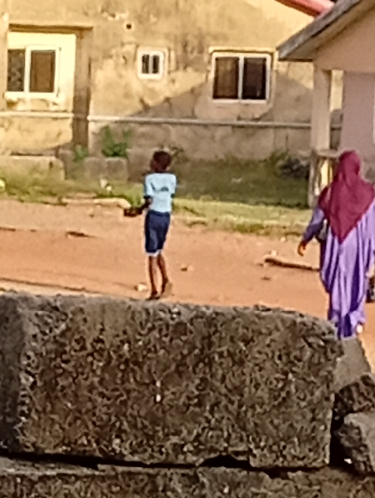A street scene in an unidentified urban location shows two people, a young boy, and a woman. The boy, wearing a light blue shirt and dark shorts, is walking toward the left. A woman clad in a long purple dress and a burgundy hijab is walking right. A large, dark-colored rock-like object is in the foreground. Behind the boy and woman are buildings with windows. The ground appears to be a dirt or sandy surface.