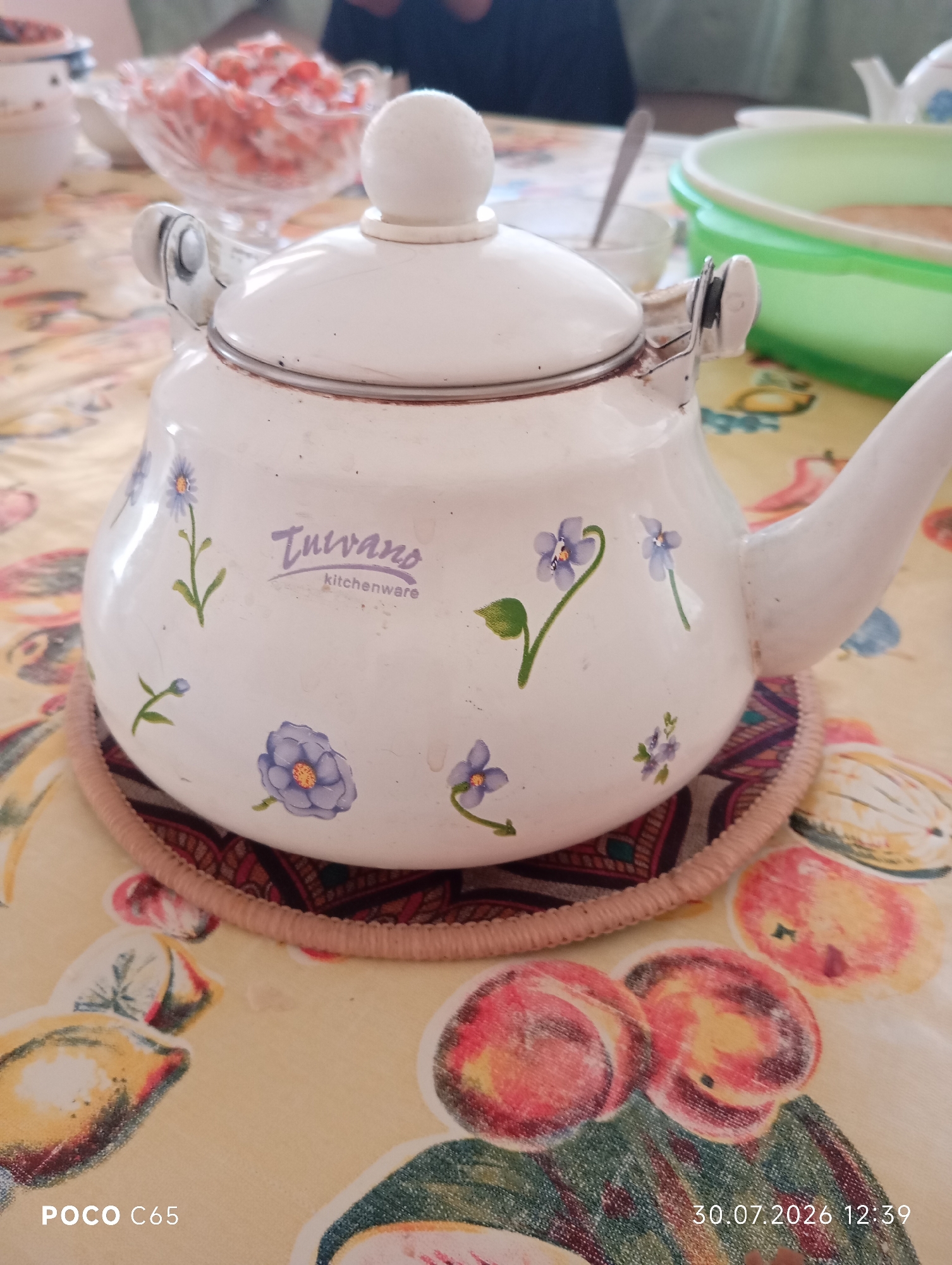 A white enameled teapot decorated with small blue flowers is placed on a patterned circular mat on a table covered with a yellow tablecloth featuring fruit illustrations. The teapot has a rounded body, a lid with a knob, and a spout. A brand name "Tulvano kitchenware" is visible on the side of the teapot. In the background, there is a glass bowl filled with a pink salad and other tableware. The scene appears to be an indoor setting, possibly a dining table. The bottom left corner of the image shows "POCO C65" and the bottom right corner shows "30.07.2026 12:39". This image is not geotagged to Beruniy, Uzbekistan.