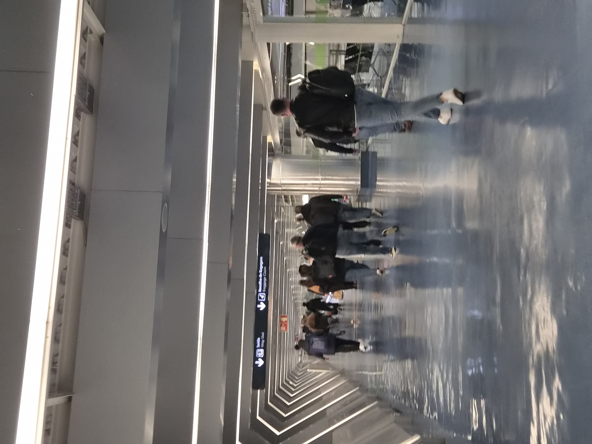 The image depicts an interior walkway at an airport. A group of people are walking along a shiny, light-colored floor. The walkway is illuminated by linear light fixtures running parallel to the walls and ceiling. The walls and ceiling are gray. There's a sign hanging on the left side with arrows and the text "Baggage Claim". The location appears to be inside Francisco Sá Carneiro Airport, located in Matosinhos, Portugal.