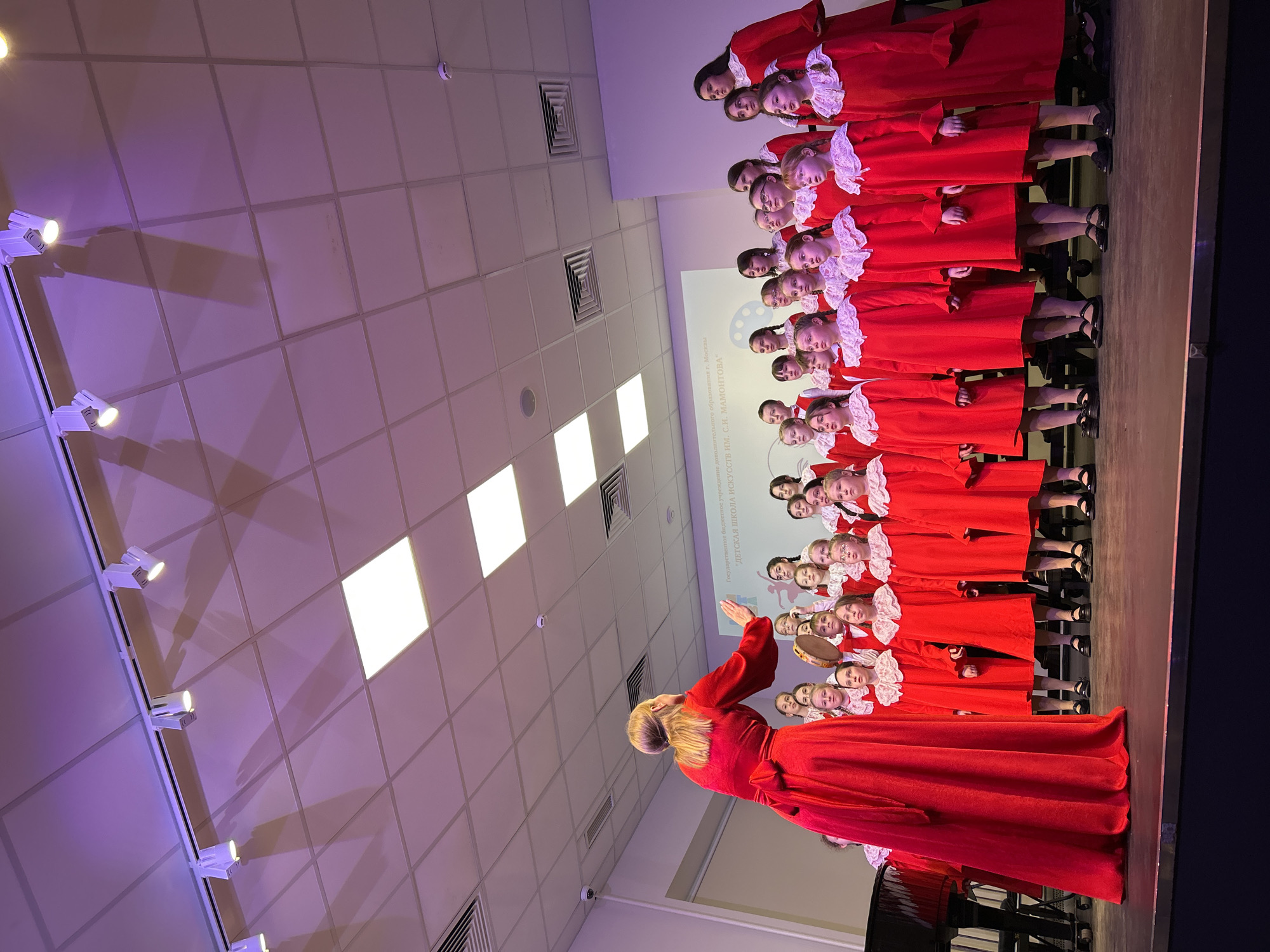 An indoor performance of a children's choir is captured in an auditorium or hall. Approximately 20-25 young girls, uniformly dressed in long red gowns with white ruffled collars and cuffs, stand in rows on a stage. Their attention is directed towards an adult woman, also in a red gown, who stands before them with her arm raised, actively conducting the choir. She has blonde hair tied back. One girl within the choir holds a tambourine.
Behind the choir, a large screen or projection displays Cyrillic text, which reads: "Государственное бюджетное учреждение дополнительного образования г. Москвы 'ДЕТСКАЯ ШКОЛА ИСКУССТВ ИМ. С.И. МАМОНТОВА'". This translates to "State Budgetary Institution of Additional Education of Moscow 'CHILDREN'S SCHOOL OF ARTS NAMED AFTER S.I. MAMONTOV'", indicating the event is associated with a children's arts school in Moscow, Russia.
The ceiling features square acoustic tiles with several bright track lights casting shadows, as well as larger rectangular recessed lights. Air vents are also visible on the ceiling. The artificial lighting, a mix of bright white spotlights and a purple/blue ambient glow, suggests an evening event or a performance in a controlled indoor environment. The children and the conductor appear focused and engaged in the musical presentation.