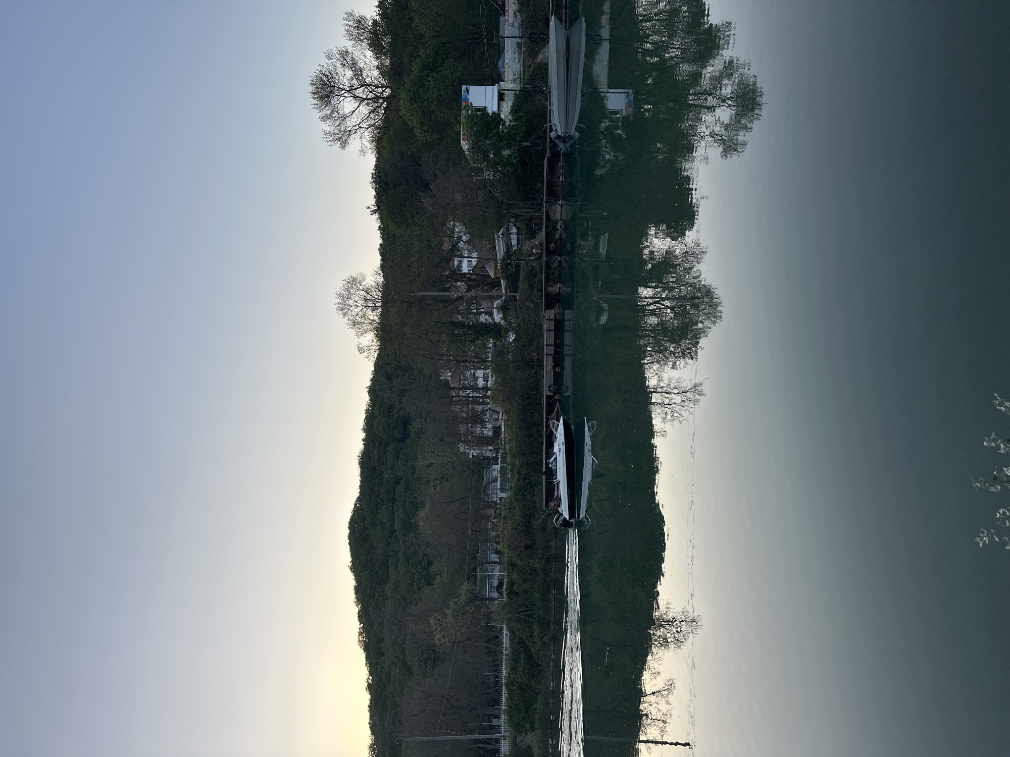The image shows a landscape scene with a body of water in the foreground, reflecting a landmass with trees and buildings. The sky is visible in the upper portion of the frame. There are boats docked along the shoreline. Due to the reflection, the image appears symmetrical along a horizontal axis. Without additional information, it is impossible to confirm the location as Novomikhaylovsky, Russia.