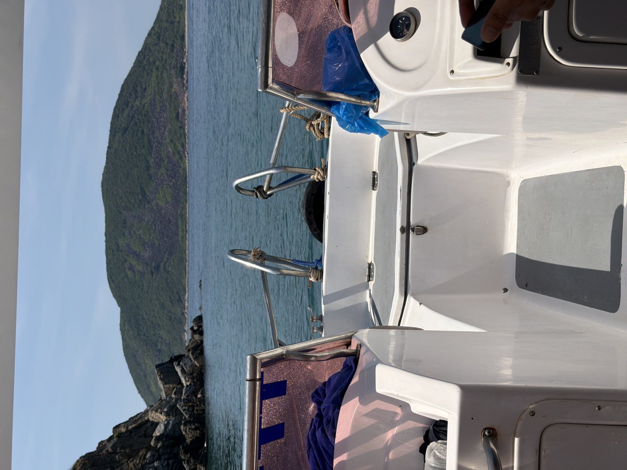 The image shows a view from a boat on the water near Bac Nha Trang Ward, Vietnam. An island or peninsula covered in green vegetation is visible in the distance on the left side of the frame. The boat's features, including the steering console, metal railings, and various components, are prominent in the foreground. A hand holding an object is visible in the top right corner. The sky is clear and blue. The sea is visible in the background. Rocks are visible near the coastline.