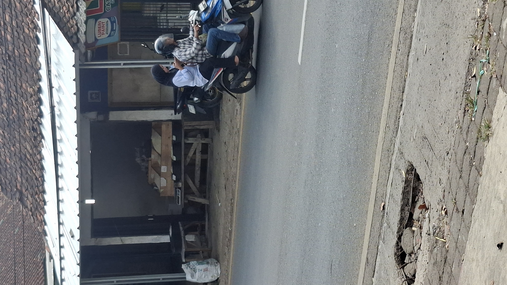 On a street in Cilaku, Indonesia, a woman in a light blue shirt and black headscarf is riding on the back of a scooter driven by a man in a plaid shirt and blue jeans. The woman is looking at her phone. They are traveling on a paved road with a broken concrete edge on the left side of the image. In the background, there is a building with a tiled roof and an open-fronted shop displaying wooden crates. A white bag is visible near the shop entrance. The environment appears to be a commercial or residential area.