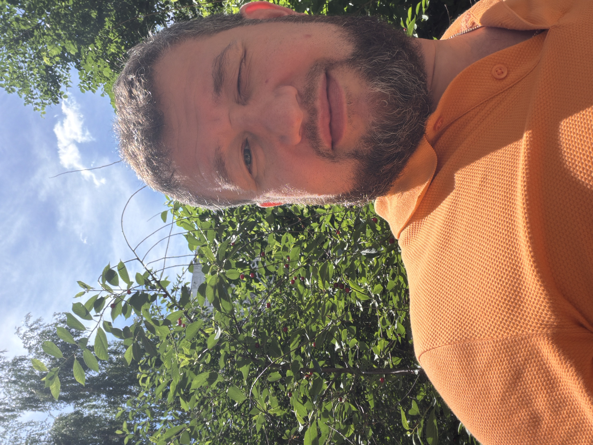 The image is a selfie taken outdoors, likely in a park or garden, given the presence of lush green foliage and a bright blue sky with scattered clouds. A man with a beard and short, light brown hair is the primary subject. He is wearing an orange polo shirt and winks at the camera. The background consists of dense green leaves, some of which appear to be from a cherry tree, with a few red cherries visible. The lighting suggests it is daytime, with sunlight casting shadows on the man's face and shirt. There is no identifiable architecture or signage to indicate a specific location within Moscow, Russia. The overall impression is that of a casual outdoor portrait.