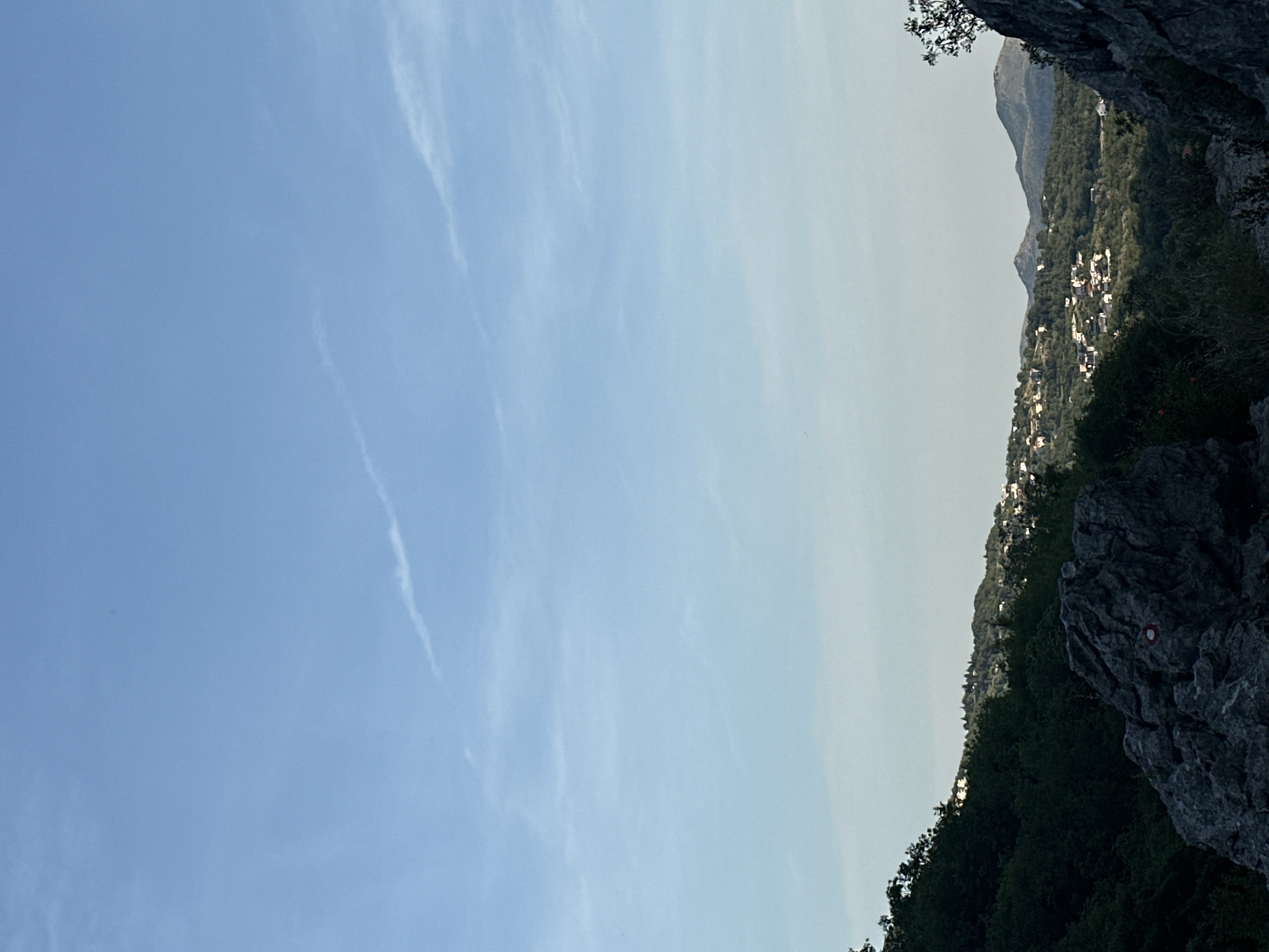 The image displays a panoramic view of a mountainous landscape under a clear, bright blue sky, taken from an elevated perspective. The sky, which occupies the upper two-thirds of the frame, shows a gradient from a deeper blue overhead to a lighter, somewhat hazy hue closer to the horizon, with subtle, wispy white cloud formations or contrails streaking across it.
In the mid-ground, a range of mountains covered in dense green vegetation stretches across the scene. A village or small settlement is visible on a hillside, characterized by numerous small white or light-colored buildings nestled among the trees. Further mountain peaks recede into the hazy distance on the right side of the image.
The foreground features rugged, dark rock formations, particularly on the right, and dense dark green foliage, suggesting a rocky and forested environment. A distinct red and white circular hiking trail marker is painted on a prominent rock face in the lower right portion of the image. The bright, even lighting suggests it is daytime. The scene captures the natural beauty of Zupci, Montenegro. No explicit text is visible, only the symbolic trail marker.
