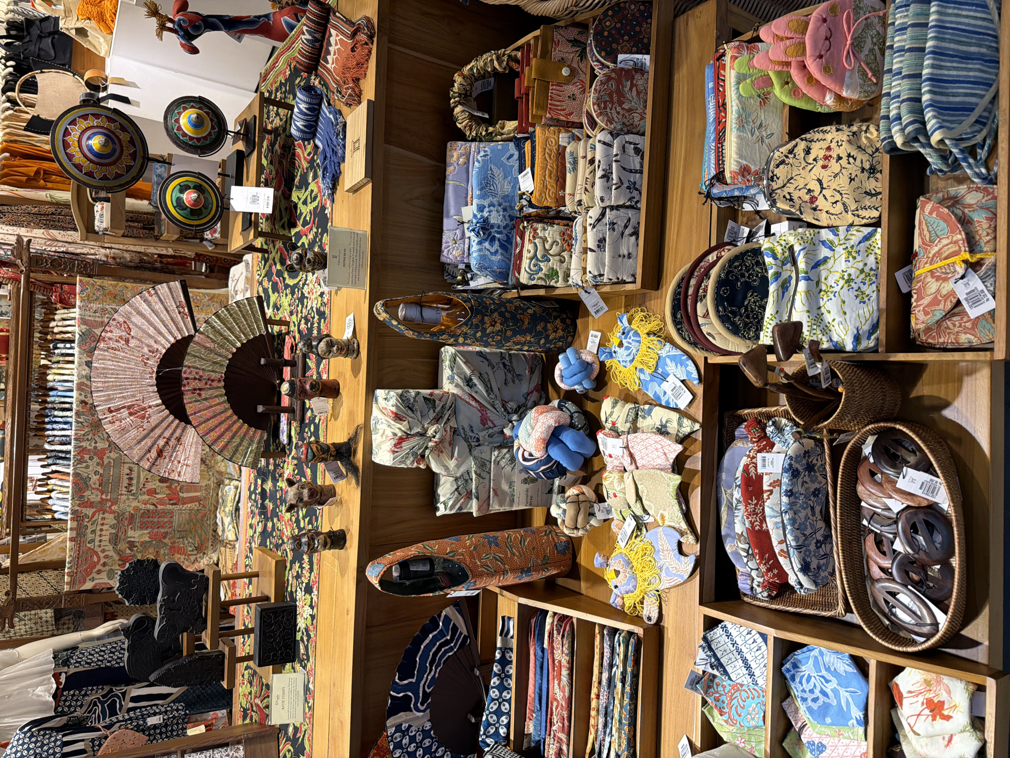 The image depicts a retail display within a shop in Kuta, Indonesia, featuring an assortment of handcrafted textiles, accessories, and decorative items. The display is organized across wooden shelves and surfaces, showcasing a variety of patterned fabrics, furoshiki wraps, small bags, and ornamental objects.
On the upper shelves, several large, decorative fans with intricate designs are displayed, along with round, colorful decorative wheels mounted on stands. A mannequin draped in patterned fabric is visible on the far left. Small carved wooden figures and decorative stamps are also present.
The mid-level shelves are filled with more fabric items, including neatly folded textiles and rolled fabrics. A prominent orange fabric bag with a blue floral pattern, designed to hold a bottle, is positioned centrally. Various small pouches and wallets made from patterned fabric are arranged in tiered wooden boxes.
The lower section of the display features a woven basket filled with dark, carved wooden objects, possibly hair accessories or decorative clasps. Another basket contains additional fabric items, and on the far right, several plush, animal-shaped items, resembling rabbits, are visible. Folded textiles with striped patterns are stacked on a shelf at the bottom right.
The overall impression is one of a vibrant marketplace or boutique offering artisanal Indonesian crafts. The lighting suggests an indoor setting, likely a well-lit shop. No people are visible in the image.
