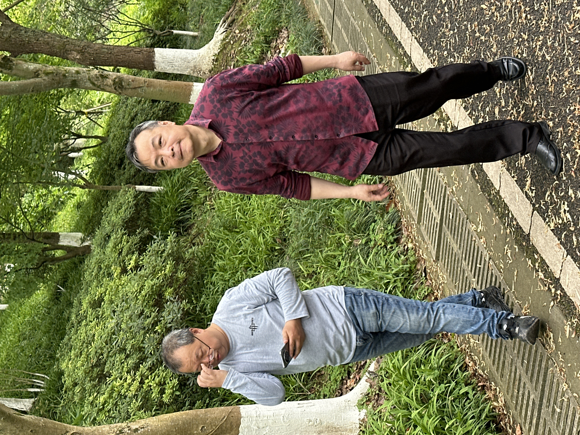 Two Asian men stand in a park-like setting. The man on the top wears a dark floral-patterned long-sleeve shirt and dark pants with black shoes. He faces the camera with a slight smile. The man on the bottom wears a light gray long-sleeve shirt, jeans, and black sneakers. He holds a phone and appears to be adjusting his glasses. The background features trees with white painted trunks, green vegetation, and a concrete walkway. The setting likely is in a park in Liangjiang, China.