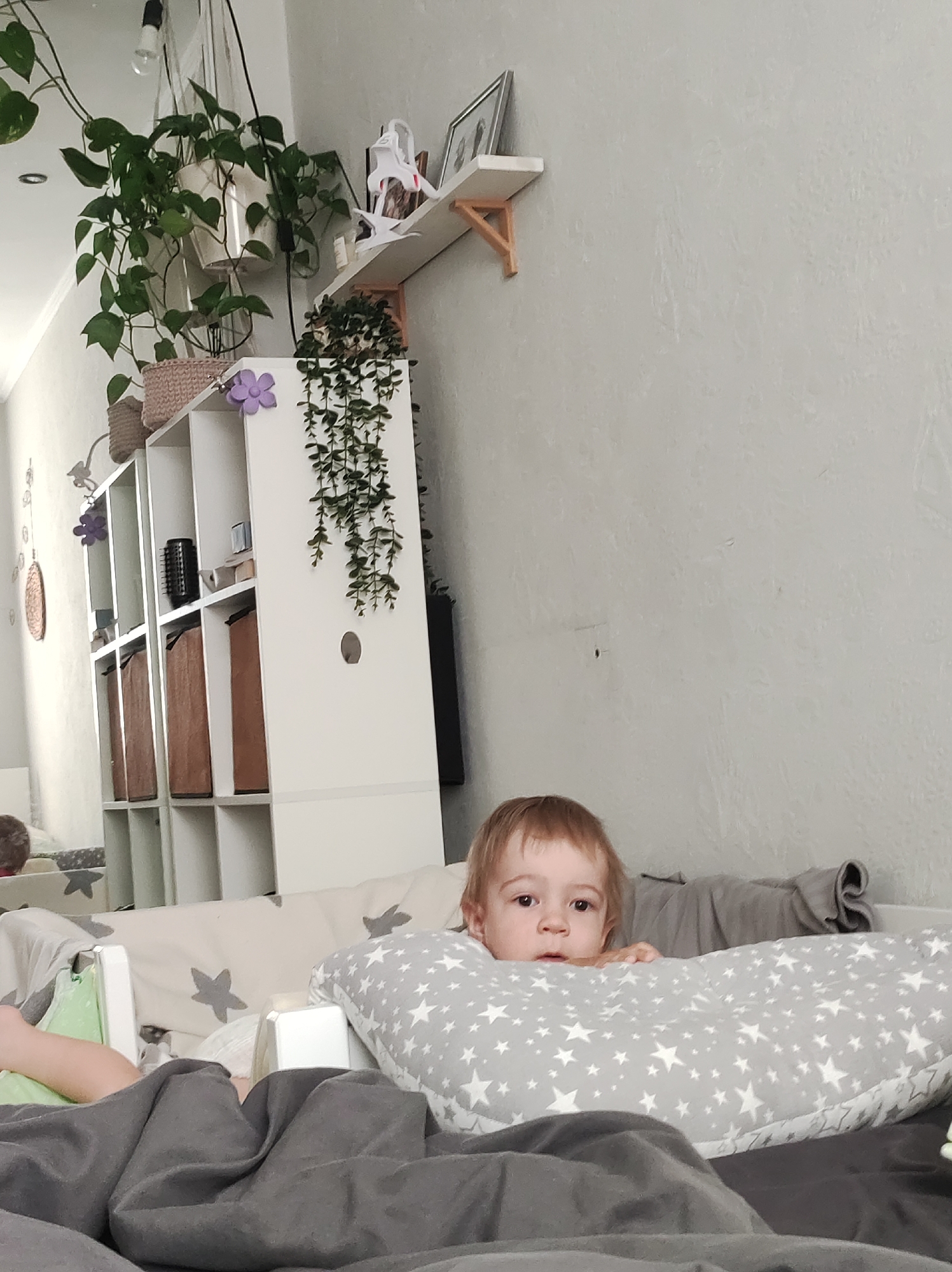 A young child with light brown hair is partially visible in a room, peering over a pillow patterned with white stars. The child's face is directed toward the camera. A gray blanket covers the lower portion of the frame. Behind the child is a crib. In the background, a white shelving unit is visible, decorated with plants and a shelf with a picture frame on it. The room has light gray walls. The image was likely taken in an indoor setting, the city location Chaikovsky, Russia is based on information available during training, but not visually verified in the photograph.