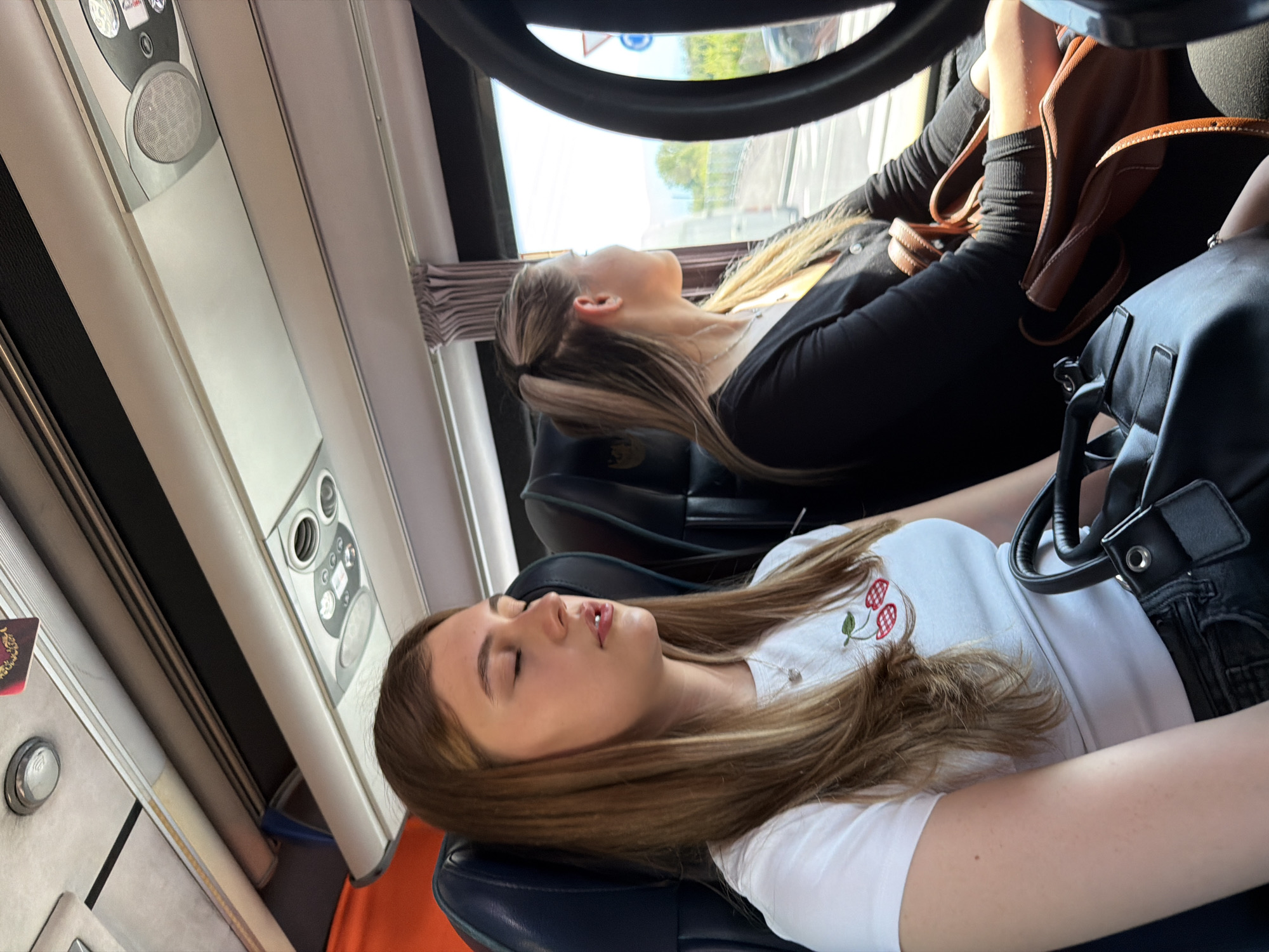 Two young women are seen resting or sleeping inside what appears to be a bus or coach, captured from a slightly elevated, angled perspective. Bright daylight streams in through the windows, indicating it is daytime.
In the foreground, a young woman with long brown hair, possibly with lighter streaks, is leaning back in her seat, seemingly asleep with her mouth slightly ajar. She wears a white t-shirt with a small red and green cherry embroidery on the chest, and black jeans. A silver necklace with a small charm is visible around her neck. A black handbag rests on her lap. Her seat is dark blue or black.
Further back and slightly to the right, another young woman with light brown or blonde hair, tied back, also appears to be asleep or resting. She wears a black long-sleeved top or jacket. Her feet, clad in black ribbed socks and brown loafers, are visible resting on what appears to be a dashboard or partition. A brown leather bag with a strap is placed near her feet.
The interior of the vehicle features overhead panels with functional buttons, speakers, and vents. On the panel above the foreground subject, text like "Call" and "Stop" is discernible near buttons. Curtains are pulled back on the window beside the woman in the background, revealing a blurred outdoor scene with trees and possibly buildings, suggesting travel through an urban or suburban area. The overall atmosphere is one of peaceful transit. The location context indicates this is in Plovdiv, Bulgaria.