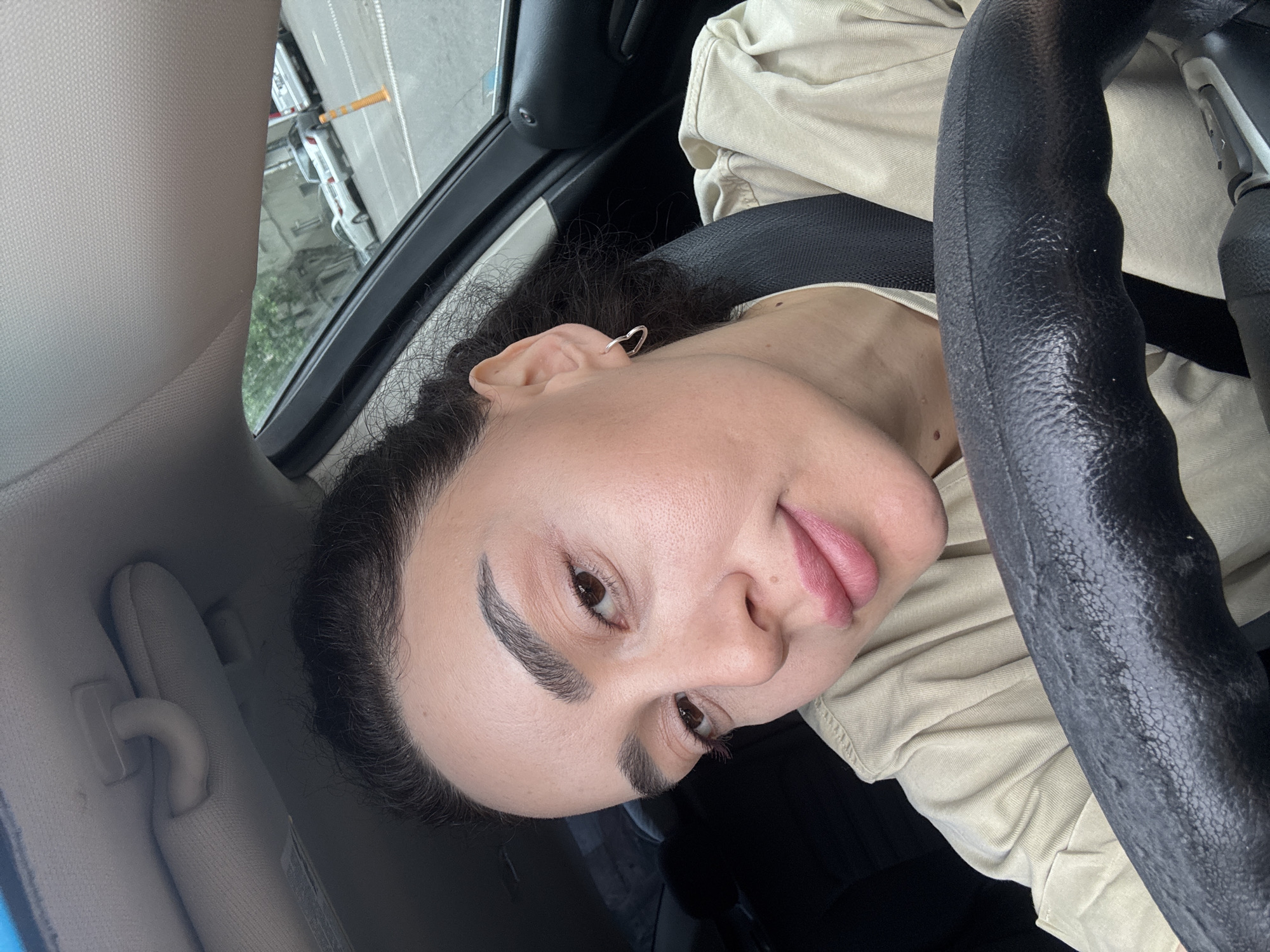 A woman with dark curly hair is in a car. She is wearing a beige shirt and a seatbelt. Her face is turned towards the camera, showing her left profile. Her tongue is slightly sticking out. She has an earring in her left ear. A black steering wheel is in the foreground. Through the car window, a street scene is visible, possibly in Tbilisi, Georgia. Several vehicles and a street with a yellow object on the side can be seen in the background. The car's interior also shows a sun visor.