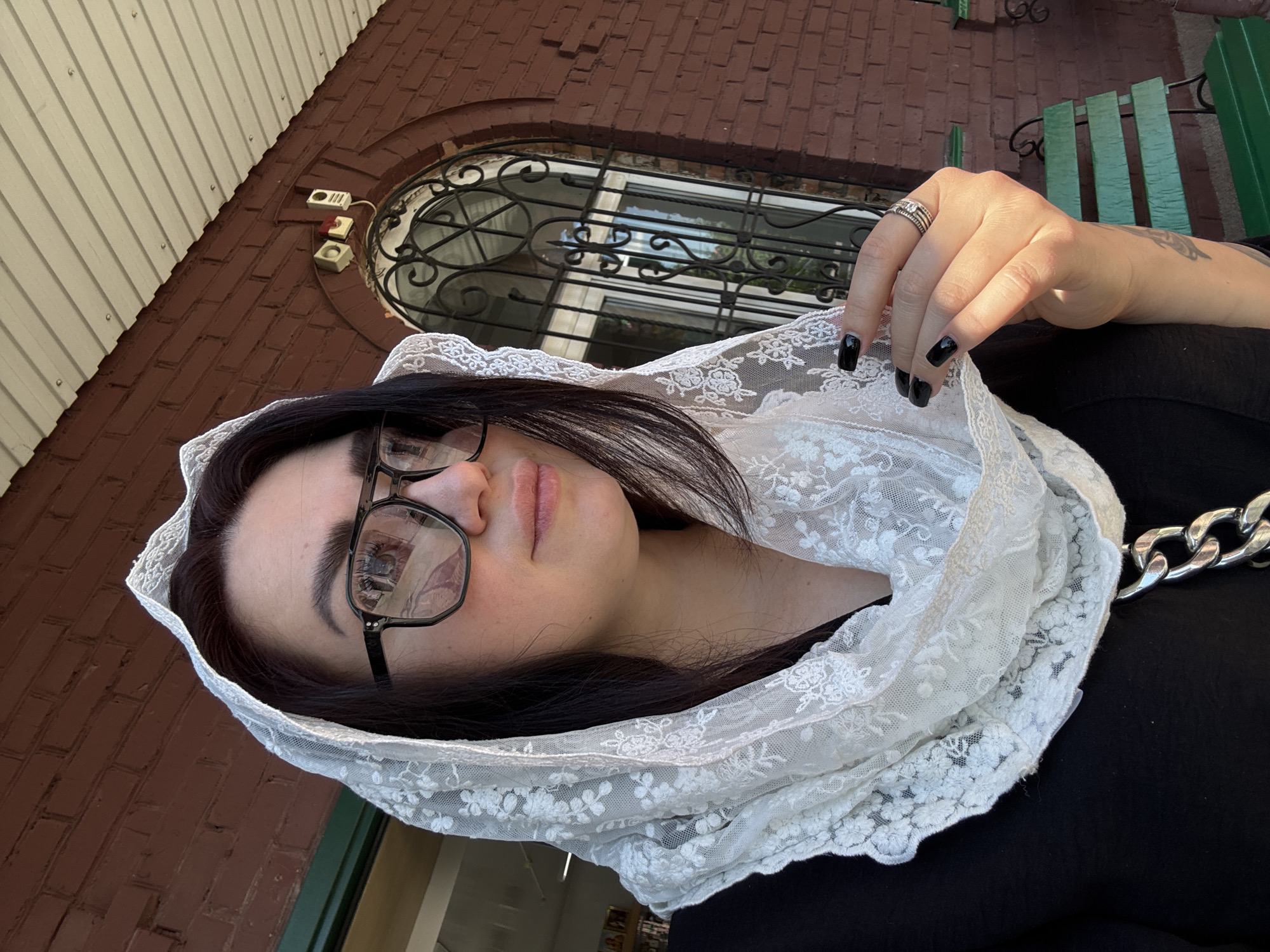 A woman wearing a black shirt and a white lace headscarf is photographed outdoors. She is looking to the right and wearing black-rimmed glasses. Her right hand is raised to her face, with black nail polish visible on her fingernails and a ring on her finger. The background consists of a brick building with a white-paneled awning and a barred window. A green bench is partially visible to the right of the woman. The lighting suggests it is daytime.