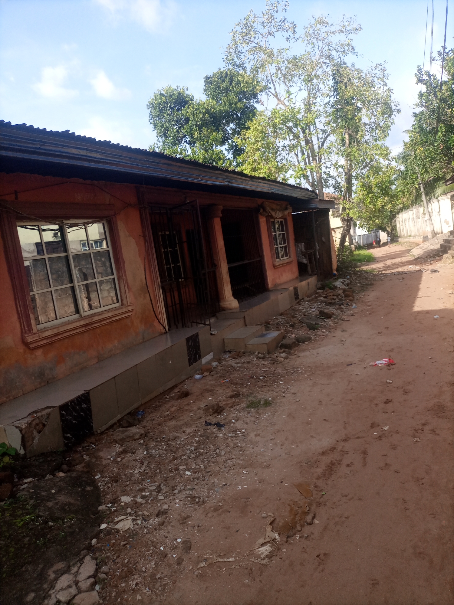The image depicts a street scene in Nkpor, Nigeria. In the foreground, a single-story building with an orange-painted facade and a corrugated metal roof is visible. The building has several windows with metal grilles and a covered porch supported by columns. The ground in front of the building is unpaved and appears to be a dirt path with scattered debris.
To the right of the building, the path continues, leading into the distance where more trees and structures are present. A white-painted wall with some decorative elements runs along the right side of the path. In the background, two figures can be seen walking away from the camera on the path. The sky above is partly cloudy with patches of blue. The overall environment suggests a semi-urban or rural setting.