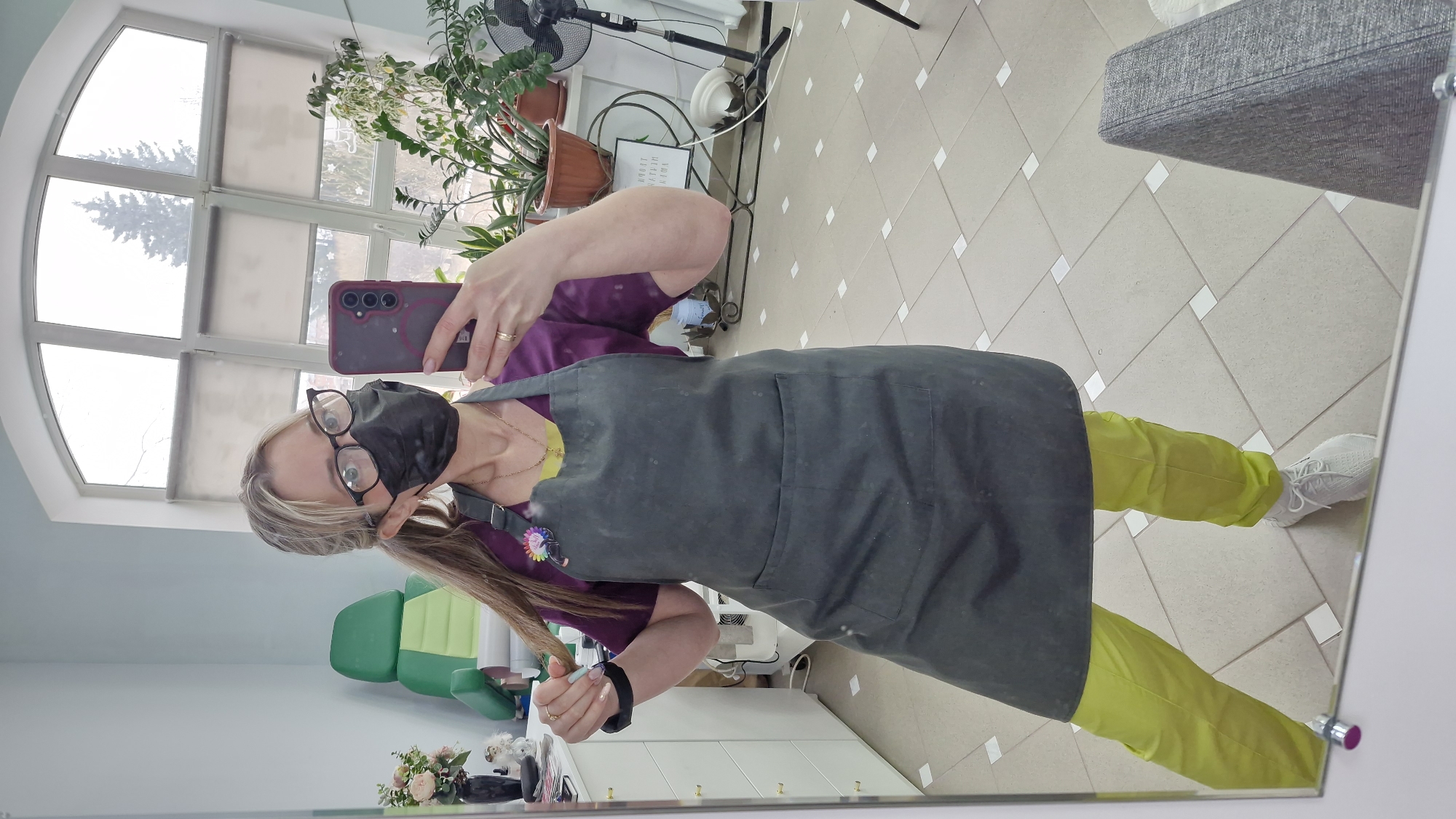 A woman in a black face mask and glasses stands in front of a mirror, holding a smartphone to take a selfie. She wears a purple shirt, yellow pants, a gray apron, and white sneakers. Her blonde hair is pulled back into a ponytail. The reflection in the mirror shows a room with a tile floor, a large arched window, and various plants on a windowsill. A green chair and a white cabinet with floral decorations are visible in the lower portion of the reflection. A fan and other equipment can be seen in the background near the window. The scene appears to be inside a salon or treatment room. There is no verifiable information about the city of Otchor, Russia in the image.