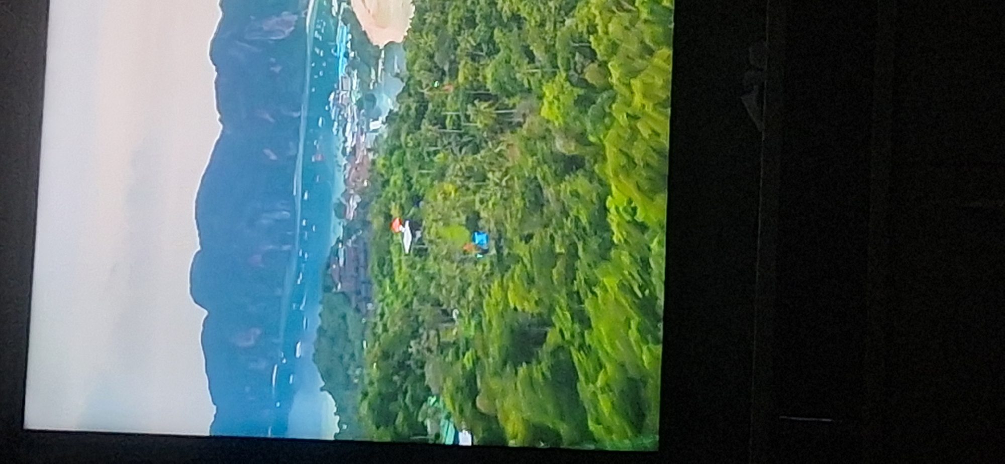 The image shows a wide aerial view of a tropical island with lush green vegetation. In the background, there are large, dark mountains and a body of water dotted with many boats. Closer to the foreground, there are buildings nestled within the trees. Two small structures are visible in the mid-ground, one with an orange roof and another with a blue canopy. The overall scene depicts a scenic island landscape. There is no discernible information about Bila Tserkva, Ukraine, in this image.
