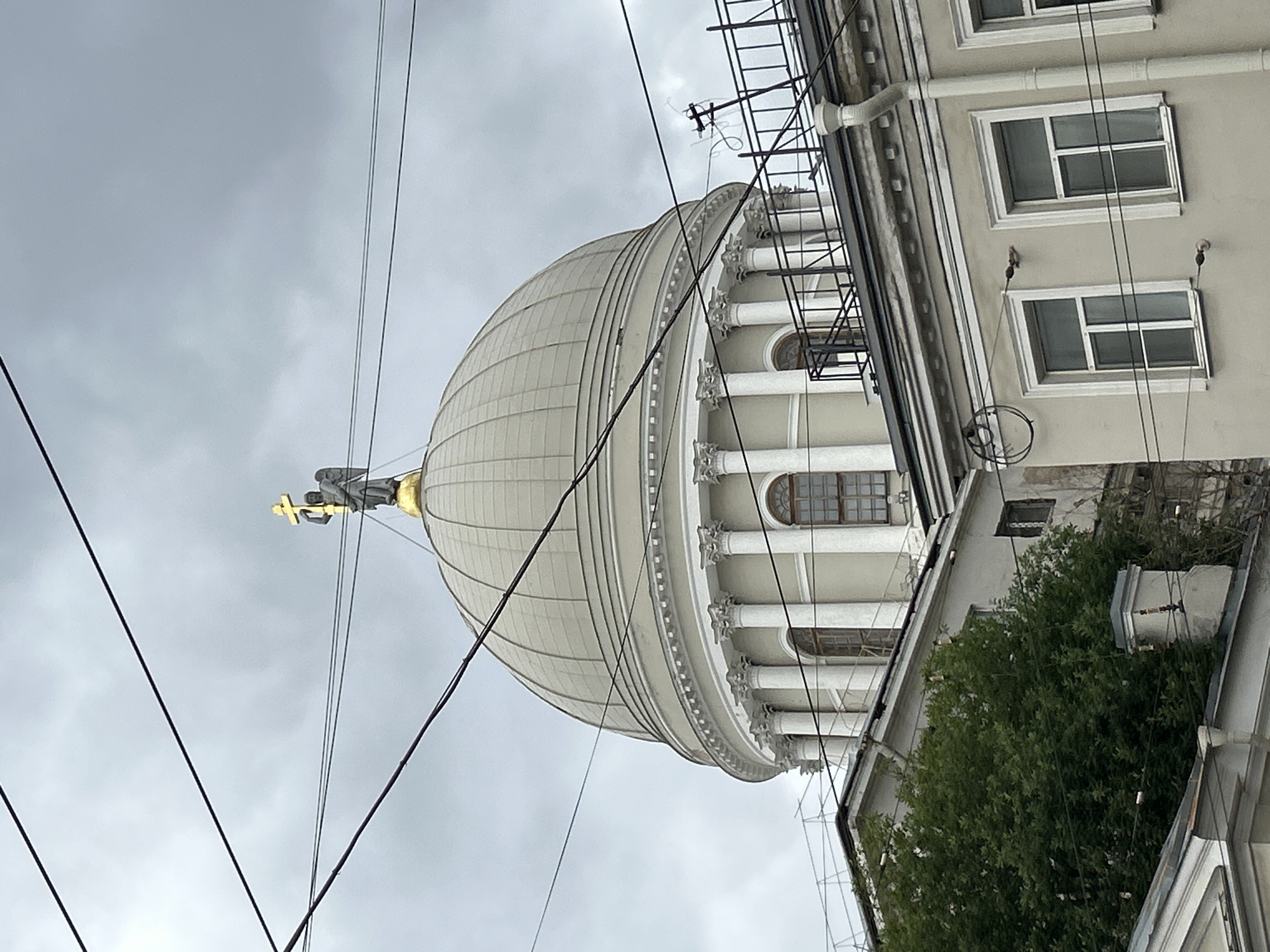 This is a photograph of an overcast sky with a large, white dome of a building prominently featured. The dome has a ribbed texture and is topped with a golden cross. An angel statue, also golden, holding the cross, is visible on top of the dome. The building has classical architectural elements, including columns and arched windows. To the right of the dome, a gray building with multiple windows is visible, along with some greenery that appears to be a large tree or bush. Electrical wires crisscross the foreground and obscure parts of the scene. The overall impression is of a grand, somewhat weathered, urban landscape under a gray sky. The orientation of the image is vertical, as if the photograph was taken looking upwards. Given the location context of Saint Petersburg, Russia, this building could be a church or a significant historical landmark. The weather appears to be cloudy, and the time of day is not clearly discernible. There is no visible text in the image.