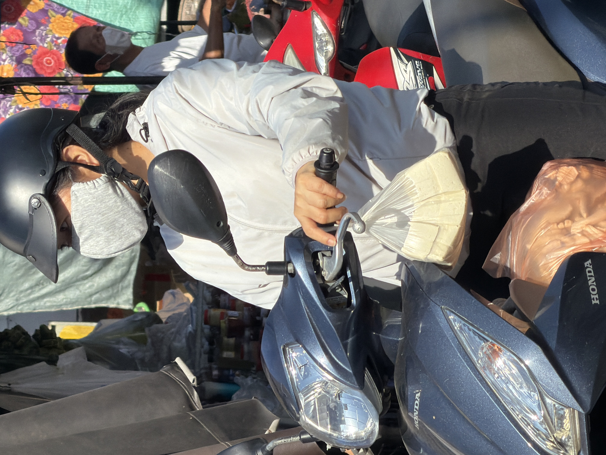 In Nha Trang, Vietnam, a person wearing a black helmet, a light gray face mask, and a light gray jacket is seated on a dark blue motorcycle, holding the handlebars. A plastic bag containing light-colored items is suspended from the handlebars, held by the person's right hand. Another person in a white shirt and white face mask is visible in the background, standing near a stall with colorful fabric coverings. The motorcycle's headlights and front fairing are prominent in the foreground. The environment appears to be an outdoor market or street scene, with various stalls and other motorcycles visible. The lighting suggests it is daytime.