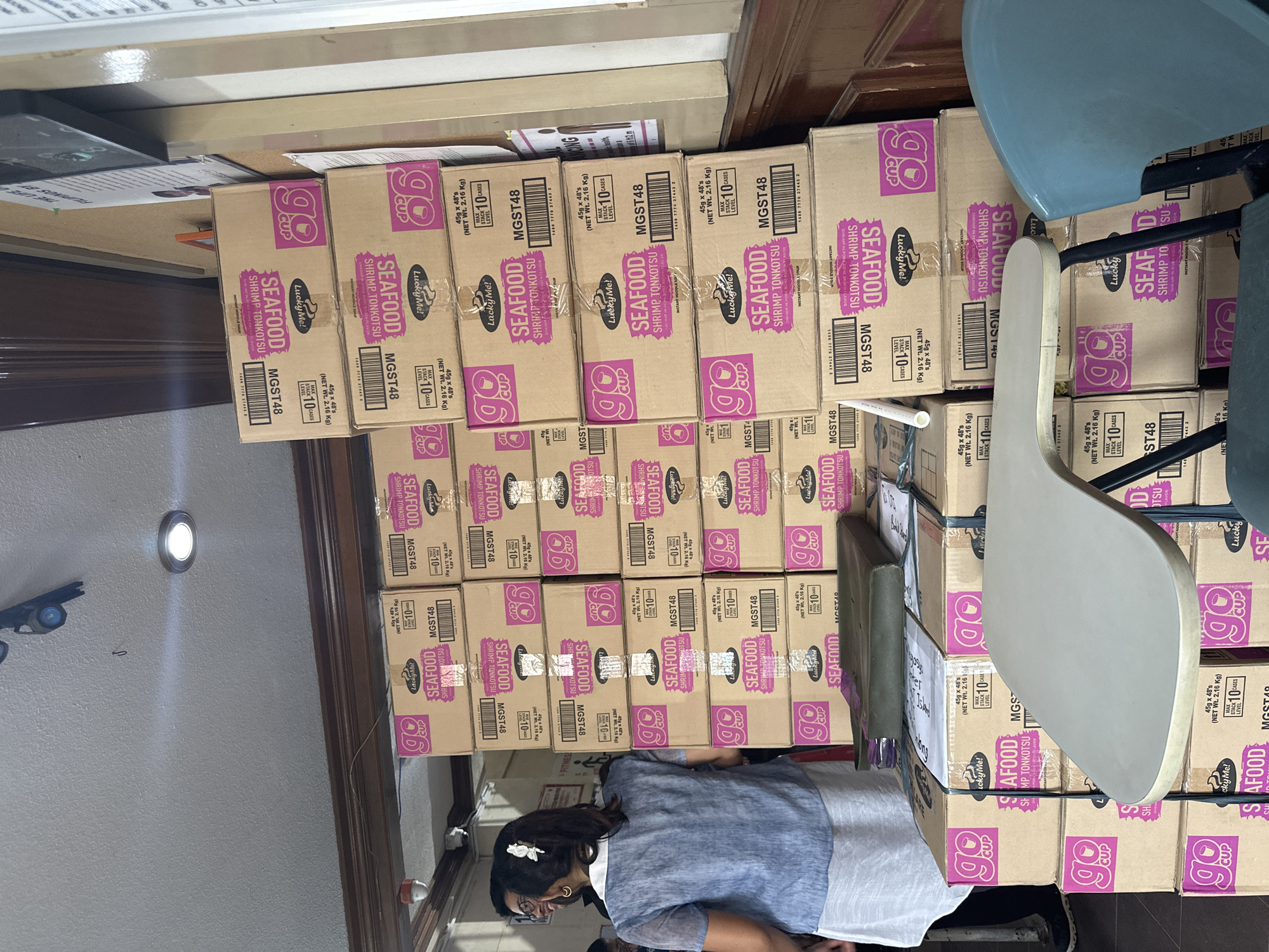 This media file depicts a storage area, likely a warehouse or stockroom, in Davao City, Philippines. The primary objects are stacks of cardboard boxes, predominantly featuring the "Lucky Me! SEAFOOD SHRIMP TONKOTSU" instant noodle brand. The boxes are neatly stacked, some reaching a considerable height. The color scheme of the packaging is a dominant pink and purple, with the "go CUP" logo prominently displayed.
A person, an adult woman, is partially visible in the lower part of the frame, facing away from the camera. She appears to be wearing a blue top and has her hair styled with a white flower. Her focus seems to be directed towards the boxes. The setting suggests an indoor environment with wooden paneling on the right side of the image and a light-colored wall with a circular light fixture on the left. The lighting appears to be artificial, indicating it could be daytime or nighttime, with no clear indicators of the weather.
The overall mood is one of organization and inventory management. The visible text on the boxes, "Lucky Me! SEAFOOD SHRIMP TONKOTSU," along with model numbers like "MGST48" and quantity information such as "45g x 48's (NET Wt. 2.16 Kg)" and "STACK 10 CASES," provides specific details about the product being stored. The presence of stacked chairs, one blue and one light-colored, further suggests a functional, possibly commercial, space.