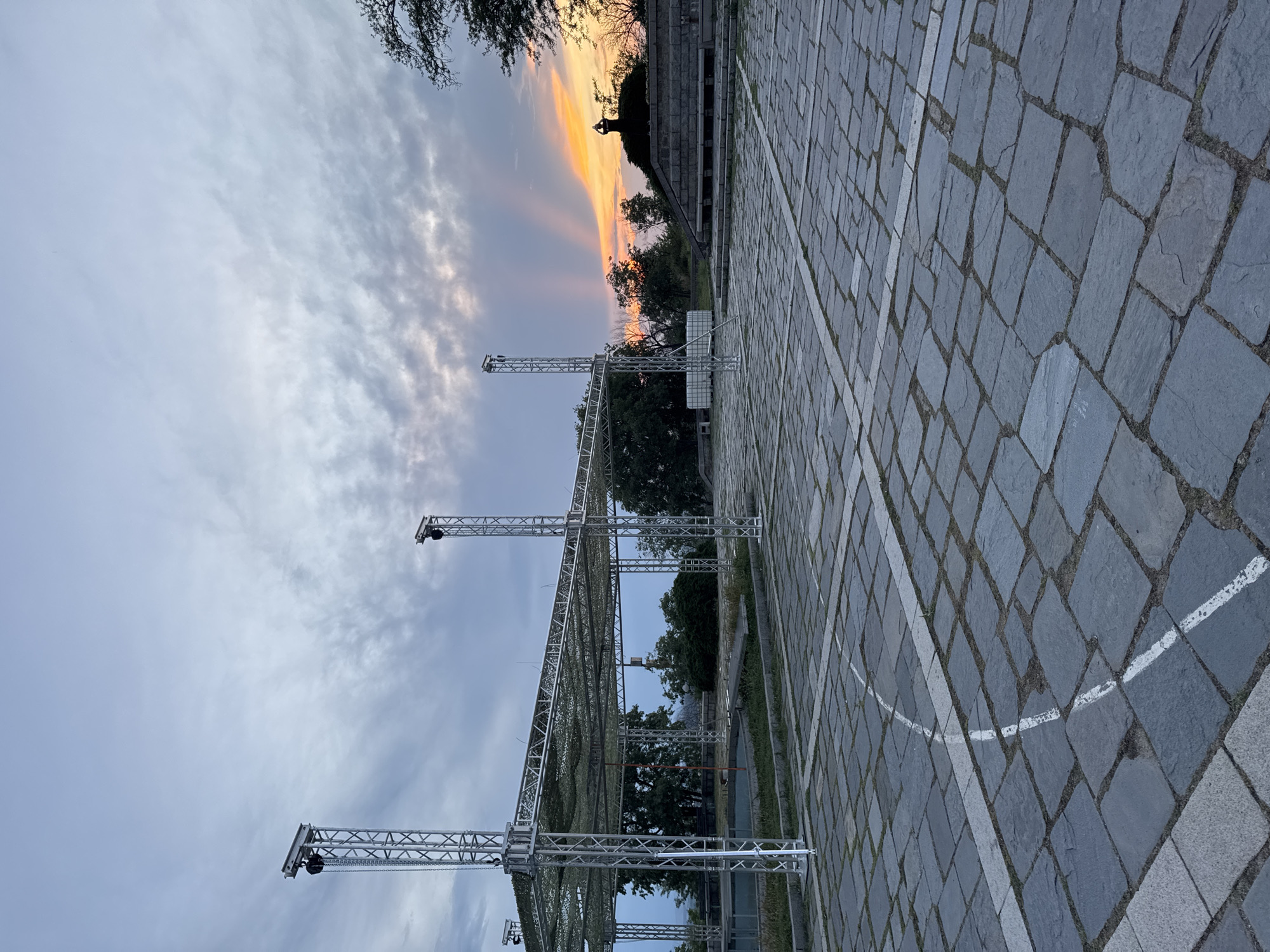 The image depicts an outdoor scene in Plovdiv, Bulgaria, captured at sunset, highlighting preparations for an event. The sky transitions from a soft blue with scattered clouds on the left to a vibrant display of orange, pink, and yellow hues on the right, indicative of dusk.
The foreground features an expansive paved area made of dark grey cobblestones, marked with white painted lines that form both straight and curved patterns, suggesting a designated space like a court or a performance area. Erected on this pavement is a large, elaborate metal truss structure, composed of interconnected aluminum segments. This rigging, likely for lights, sound equipment, or a stage, features tall vertical supports and an overhead grid, parts of which are covered with green camouflage netting. Several fixtures resembling lights or cameras are mounted on the trusses, and chains hang from some sections. A white cube-shaped container, possibly a water ballast or equipment box, is visible near one of the truss bases.
In the mid-ground, behind the truss system, green trees and foliage are visible. Further back on the right, a distinctive multi-tiered stone structure or building, characteristic of historical architecture, rises against the colorful sky. A faint silhouette, possibly a statue or a person, can be seen atop this structure. No people are visible, indicating that the image captures either the setup or teardown phase of an outdoor event. Faint, barely legible text or graffiti, including what appears to be "THE LIVE STORY Plovdiv," is inscribed on some of the cobblestones, reinforcing the context of an event in the city.