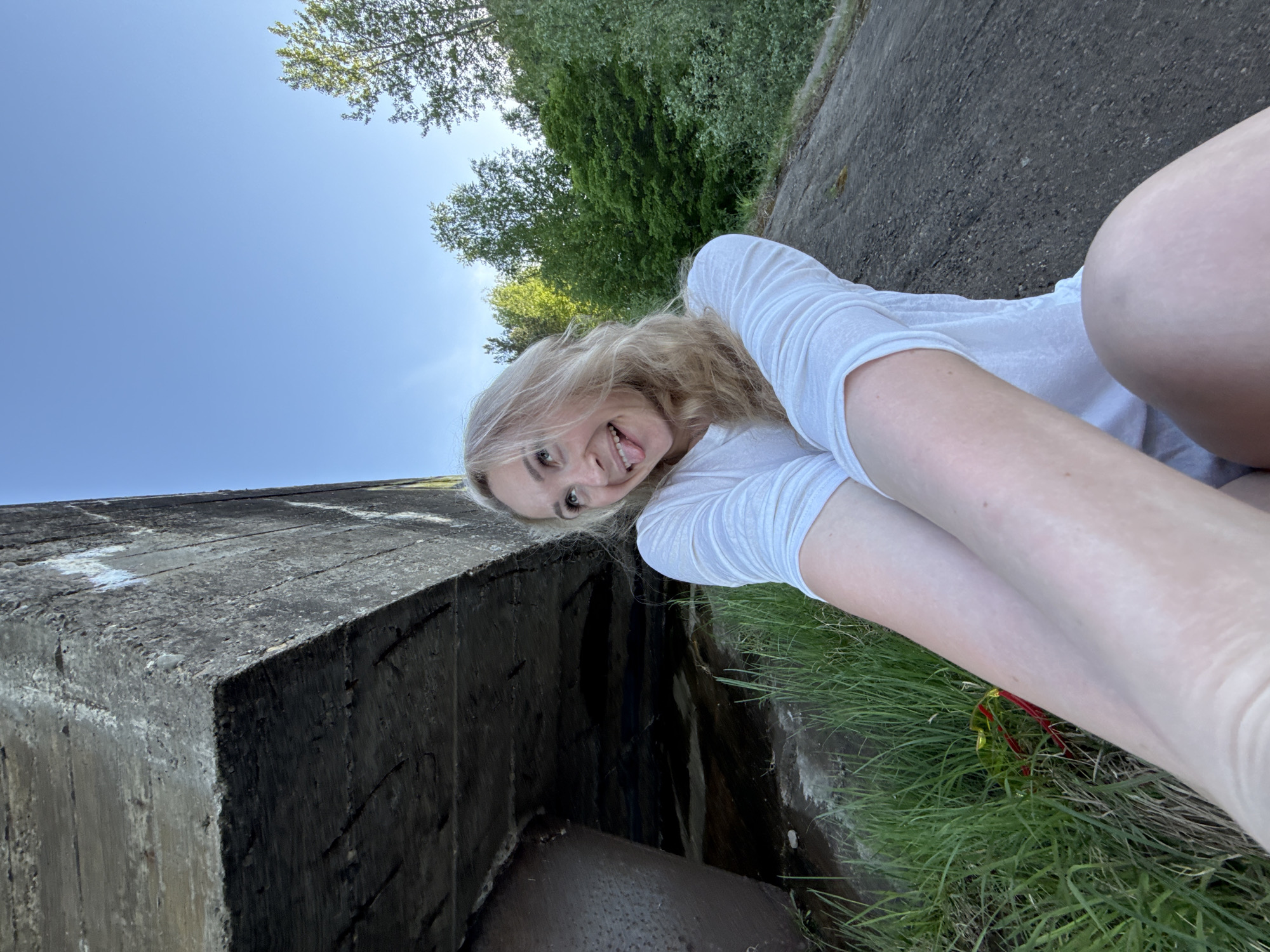 A fair-skinned young woman with blonde hair is lying on her side near a concrete structure. She is wearing a white long-sleeved shirt. Her head is tilted back, and she is sticking out her tongue with a smile. In the background, there are trees and a blue sky. The location appears to be outdoors, with grass and a concrete slab visible. There is no evidence to suggest this is in Noviy, Russia.