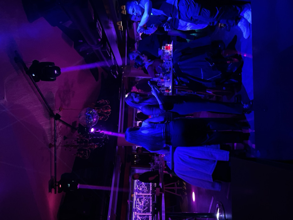 The image depicts an indoor scene in a darkly lit bar, nightclub, or lounge, characterized by vibrant artificial blue and purple lighting, indicative of an evening or nighttime event.
In the foreground, at the bottom-left, a person with long hair is seen from behind, possibly wearing a light-colored top. To their right, another individual with long hair stands near the bar counter, engrossed in their mobile phone. The bar counter stretches across the mid-ground, adorned with various bottles and glasses. Several other patrons and possibly staff members are visible standing or leaning at the far end of the bar towards the bottom-right, their features largely obscured by the dramatic colored illumination. People appear relaxed and engaged in socializing.
Overhead, in the upper-left and upper-middle sections of the frame, spotlights cast intense purple beams across the space, adding to the dynamic atmosphere. A disco ball hangs in the upper-middle, reflecting the colorful lights, and beneath it, a decorative chandelier-like fixture is visible. The overall activity suggests a social gathering where people are enjoying drinks and conversing in a lively, nocturnal environment. The setting points to a place of entertainment, possibly in Drapetsona, Greece. No clearly legible text is discernible; however, decorative graphics are present on the structures behind the bar.