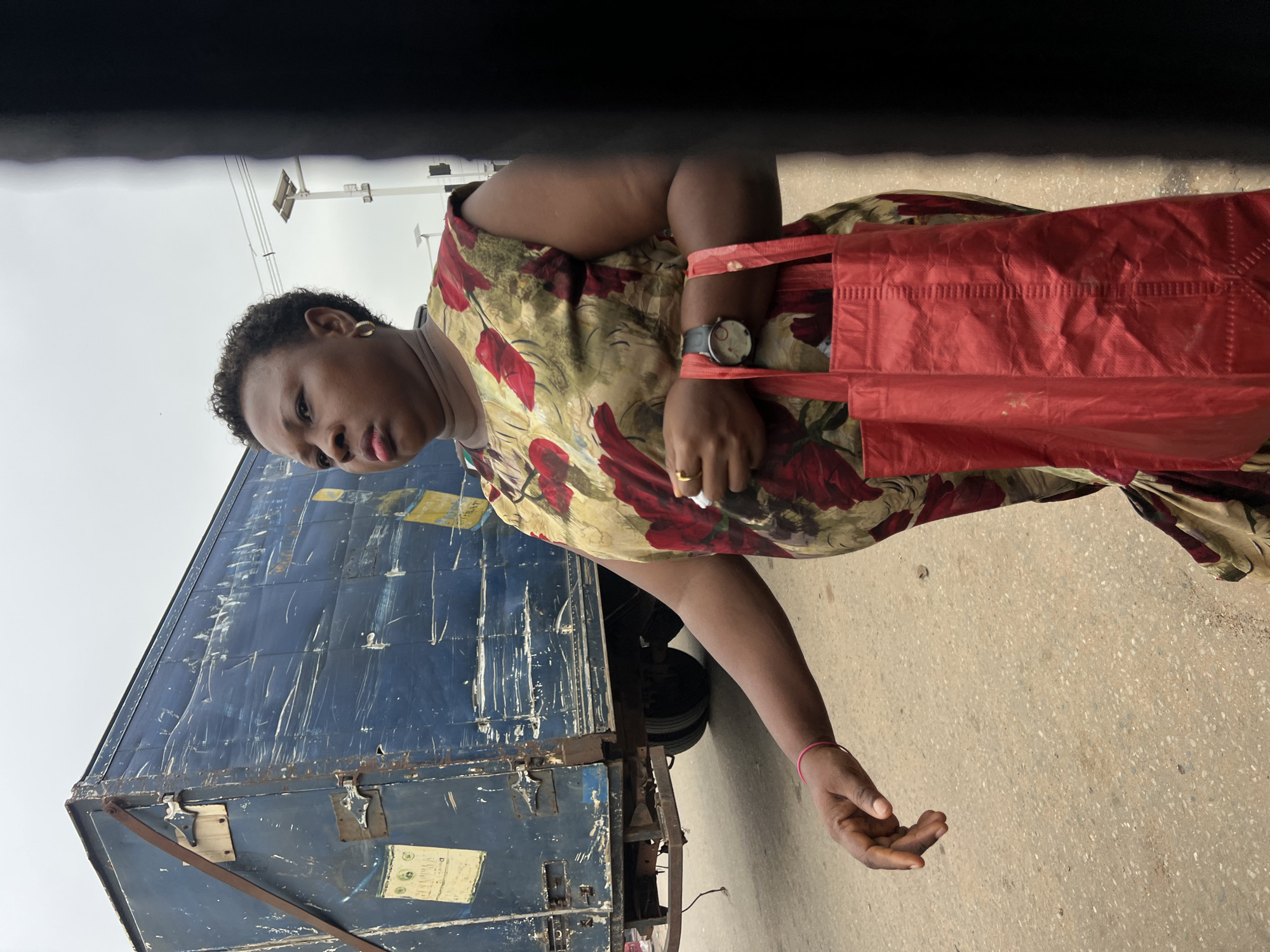 A woman stands next to a large blue truck in Nkpor, Nigeria. She is wearing a floral-patterned dress with red and green flowers and a light-colored scarf. She has a red shopping bag slung over her shoulder and a watch on her left wrist. Her right hand is extended slightly, with fingers open. The truck appears to be old and weathered, with visible scratches and rust marks. Yellow labels with text are attached to the side of the truck. The ground is paved with asphalt. The sky is overcast. The image appears to have been taken during the daytime.
