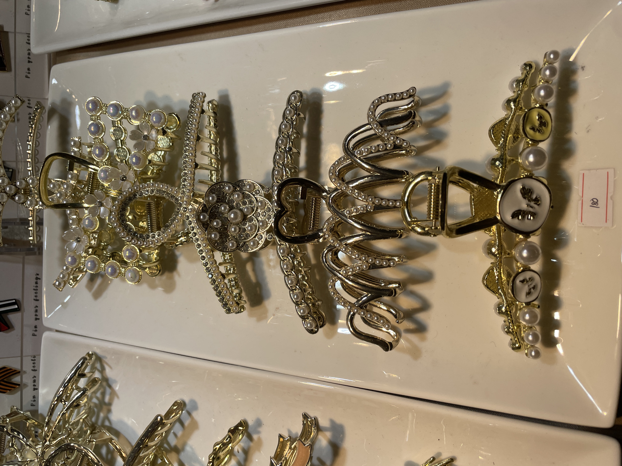 The image shows a close-up, eye-level shot of several gold-toned hair claw clips displayed on white rectangular plates. The clips are elaborately decorated with faux pearls and some feature intricate designs such as flowers, hearts, and even small enamel charms depicting butterflies. They are arranged vertically on the plates, with some overlapping. In the background, other similar hair accessories are visible, along with small cards that appear to have handwritten text and prices. The lighting suggests an indoor retail environment, possibly a stall or display case in a market or boutique. A price tag with the number "180" written on it is visible at the bottom of the image. The overall impression is one of a display of decorative hair accessories, likely for sale.
This scene is consistent with what might be found in a shopping center or market in Davao City, Philippines, such as Gaisano Mall, SM City Davao, or a smaller local boutique.