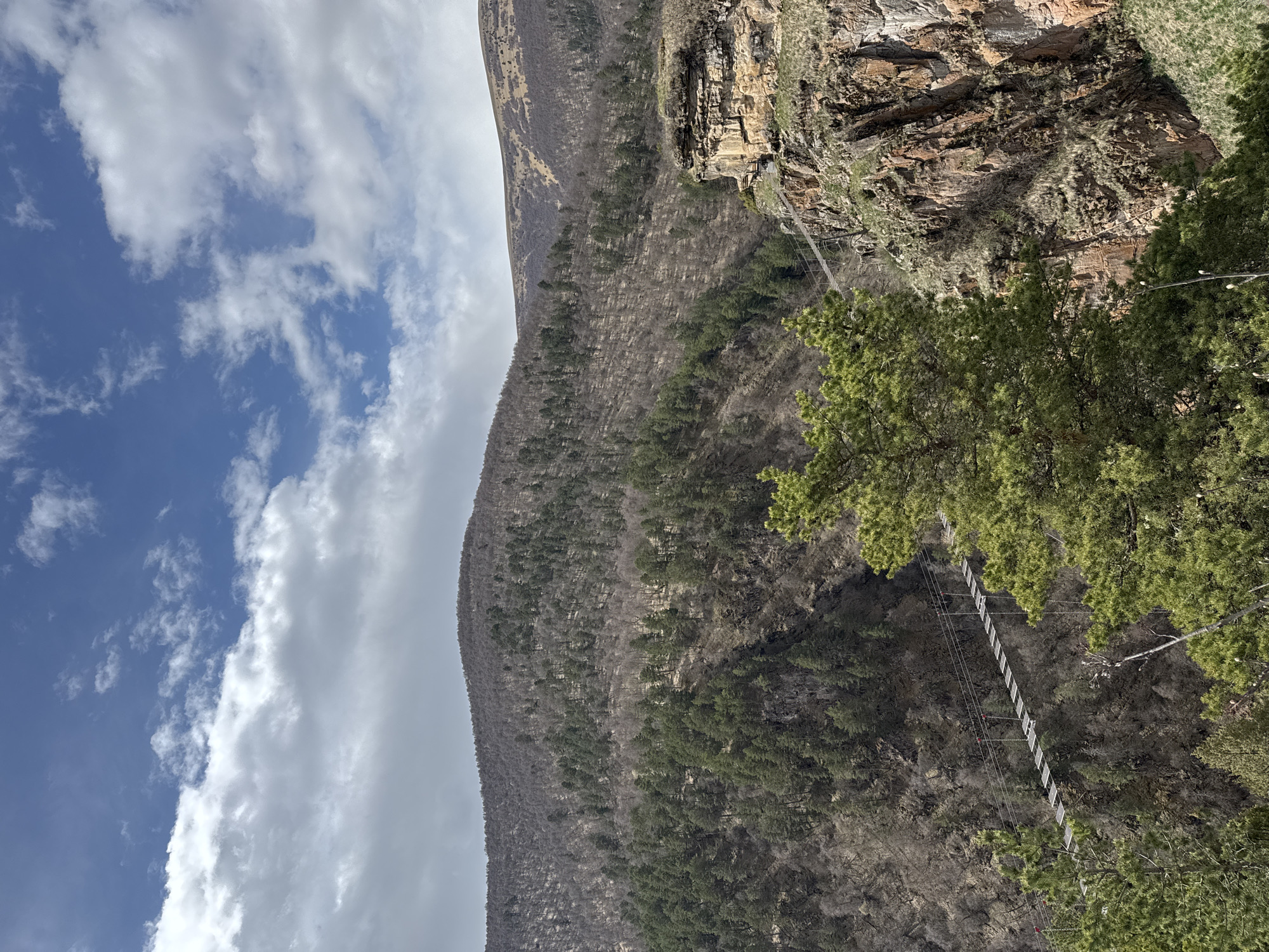 The image shows a mountainside scene in an unknown location, likely in Russia based on the prompt. The sky is partially cloudy, with blue patches interspersed with white clouds. A large mountain dominates the scene, with a mix of bare rock, trees, and vegetation covering its slopes. A suspension bridge is visible, spanning a gap between two sections of the mountain. The image is rotated.