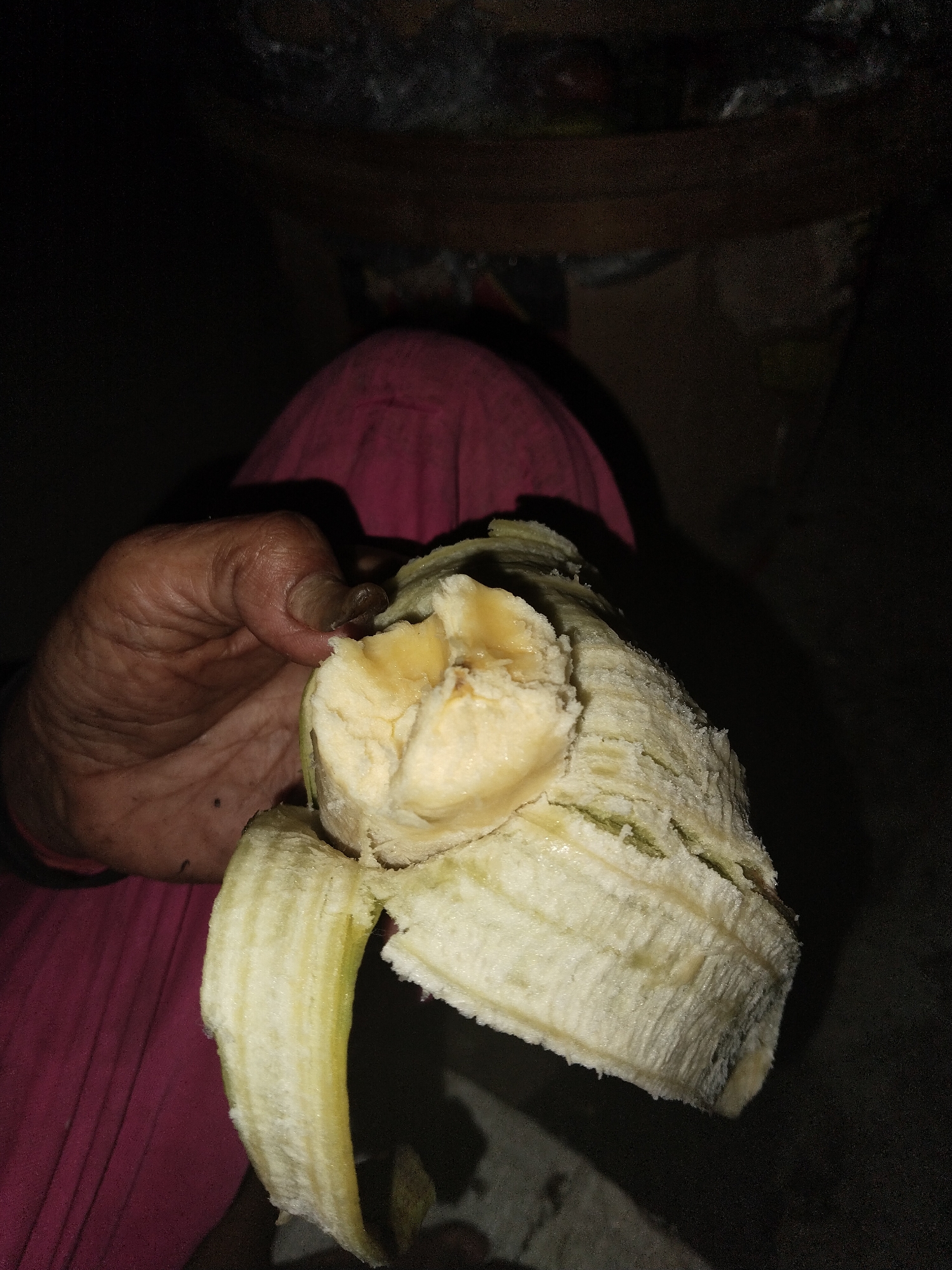 A hand holds a partially peeled, thick banana, exposing its creamy white flesh. The banana's peel is thick and fibrous with a light green tinge. The hand holding the banana appears weathered with visible wrinkles and a mole. The person holding the banana is wearing a pink top. The location and city are unknown.