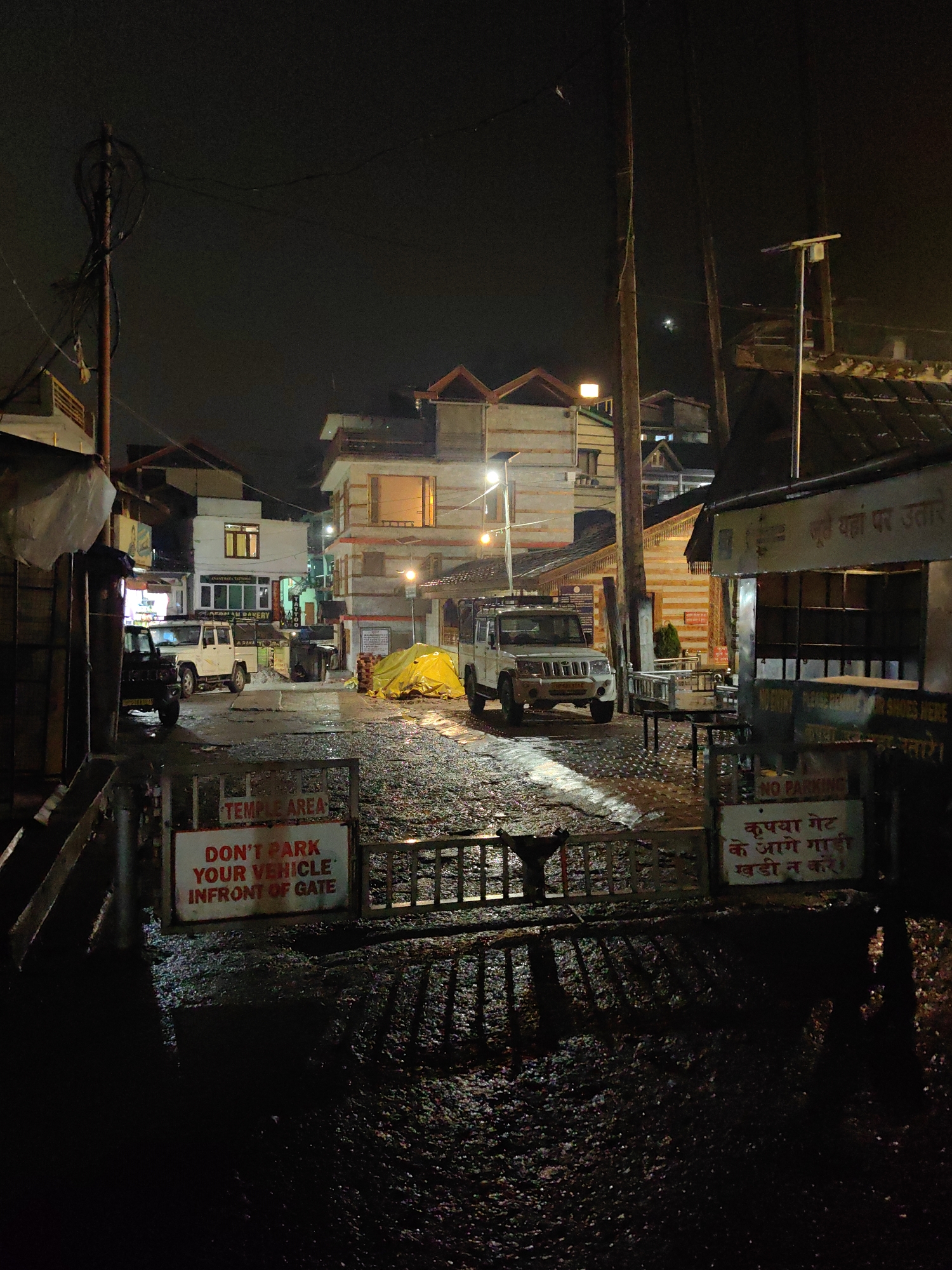 This is a night-time photograph taken in Vashisht, India, likely on a street or a parking area. The scene is dimly lit, with streetlights and lights from buildings illuminating the wet ground and the objects present. In the foreground, there is a metal gate with a sign that reads "TEMPLE AREA" and "DON'T PARK YOUR VEHICLE INFRONT OF GATE". To the right of the gate, another sign in a different script is visible. The ground appears to be paved with gravel and is wet, reflecting the available light.
Further into the scene, several vehicles are parked. Two white SUVs or similar vehicles are prominent. One is positioned in the middle of the street, and another is parked to the left. A dark-colored vehicle is parked to the far left. A large, bright yellow tarp covers a pile of something, possibly goods or construction materials, situated between the vehicles.
The background consists of multi-story buildings with traditional architecture, featuring wooden accents and sloped roofs. Some windows are lit, indicating occupancy. Electrical poles and wires are visible, typical of a town setting. The overall impression is of a quiet, possibly humid or rainy night in a populated area of Vashisht.