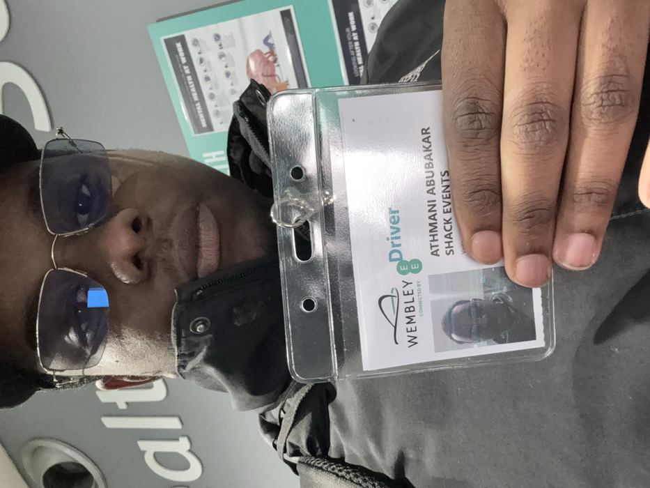 The image, rotated 90 degrees clockwise, presents a close-up portrait of a person, likely a man, with dark skin and dark hair, wearing rectangular, reflective sunglasses and a dark jacket. He is holding up an identification card in a clear plastic holder, displaying it towards the viewer with a neutral expression.
The ID card clearly identifies the individual as "ATHMANI ABUBAKAR" and indicates an affiliation with "SHACK EVENTS." It prominently displays the "WEMBLEY" logo and text that reads "Driver," along with "CONNECTED BY E" (likely referring to the EE mobile network). A small ID photo, consistent with the person in the main image, is embedded on the card. The person's fingers, with neatly trimmed nails, are visible as he holds the card.
In the background, a light blue-green poster is partially visible, containing text such as "MENTAL HEALTH AT WORK" and "LOOKING AFTER YOUR AL HEALTH AT WORK." Another partial word, "alt," is visible from what appears to be a separate sign. The indoor setting, bright lighting, and the "Wembley" detail on the ID card suggest the location is an event venue or workplace in Wembley, United Kingdom.