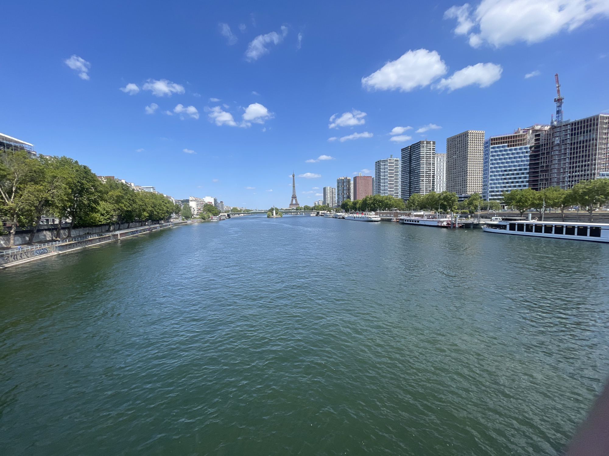 This media file is a landscape photograph taken on a clear, sunny day in Paris, France. The image depicts the Seine River flowing through the city, with a cityscape visible on both banks.
The main subject is the river itself, a wide expanse of green water with gentle ripples reflecting the bright blue sky and scattered white clouds. Several boats are moored or sailing on the river, including what appear to be tour boats or river cruises.
On the left bank, a tree-lined embankment lines the river, with a stone wall featuring graffiti. Beyond the trees, various buildings are visible, some appearing older and more traditional.
On the right bank, a modern skyline dominates the scene. Tall, contemporary buildings, including residential or office towers, are prominent, with one building under construction featuring a red crane. The Eiffel Tower is visible in the distance, a recognizable landmark of Paris, positioned over a bridge spanning the river.
The time of day appears to be midday, given the bright sunlight and the shadows. The weather is fair and pleasant, indicated by the clear blue sky and scattered clouds. The overall mood is one of urban tranquility and scenic beauty. No people are prominently featured, and the focus is on the architectural and natural elements of the Parisian landscape. There is visible text on the graffiti on the wall along the left bank, but it is not clearly legible.
