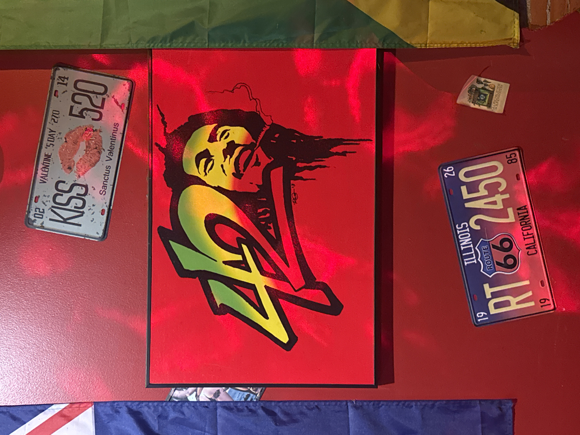 A red wall is decorated with several items. Centered is a square red canvas featuring a portrait of a figure reminiscent of Bob Marley, rendered in yellow, green, and black with bold strokes. To the left of the canvas is a license plate with the words "KISS" and "Sanctus Valentinus" along with the number "520". To the right is another license plate marked "ILLINOIS" and "CALIFORNIA," with the number "2450" and the "Route 66" logo. Above the license plate on the right is a small, faded paper item with illegible illustrations. Above the scene are flags colored green and yellow. Below the license plate are blue and white banners. Đà Nẵng, Vietnam is not evident in the image.