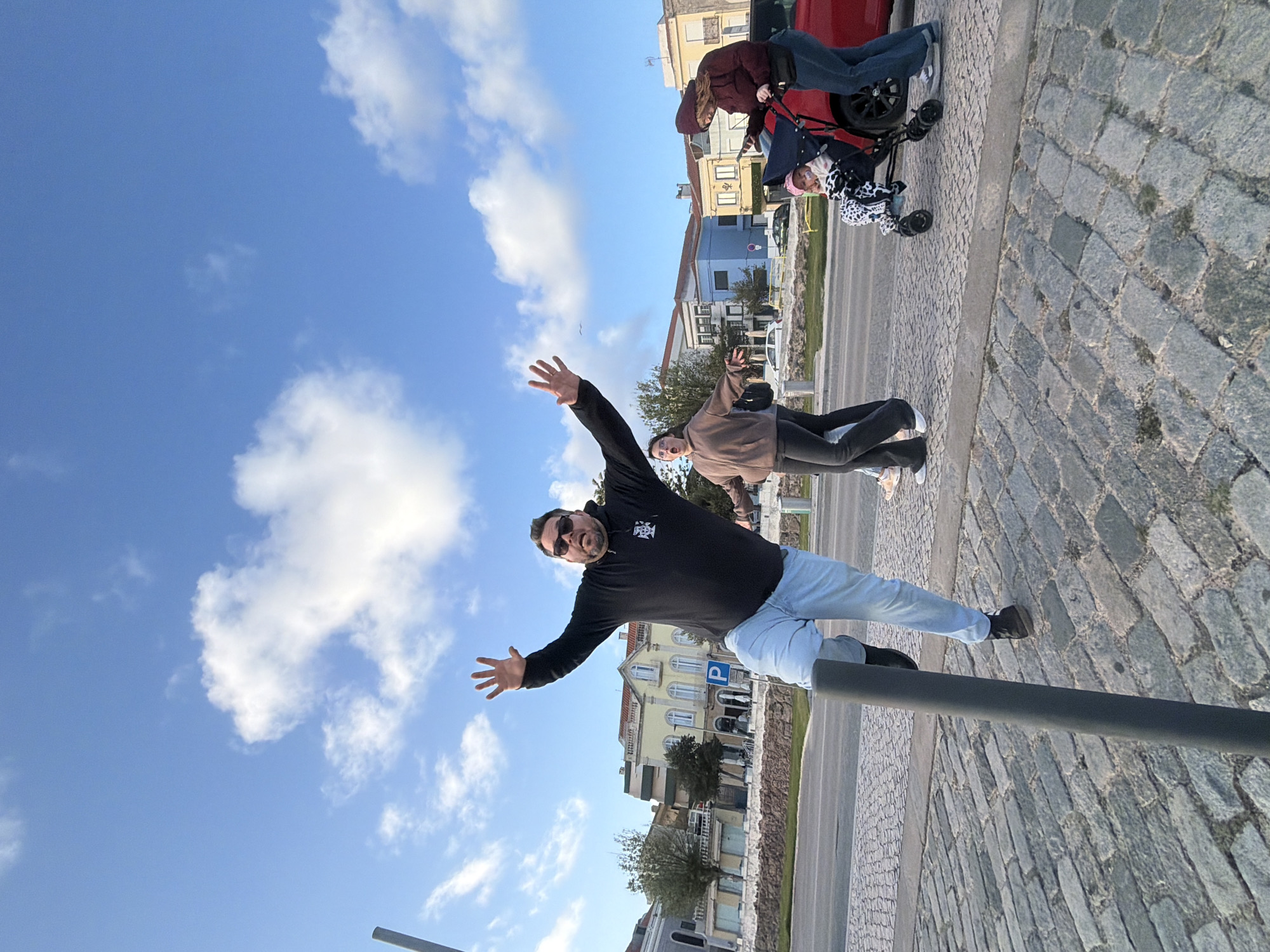 The image shows an outdoor scene in Figueira da Foz, Portugal, featuring a man in a black hoodie, light blue jeans, and sunglasses, standing on a gray metal post. He has his arms outstretched. Behind him, a woman in a brown jacket and black pants is posing with her arm raised. In the background, there are parked cars, a person pushing a stroller, and buildings, including a yellow building with a "P" parking sign. The ground is paved with cobblestones, and the sky is blue with scattered clouds.