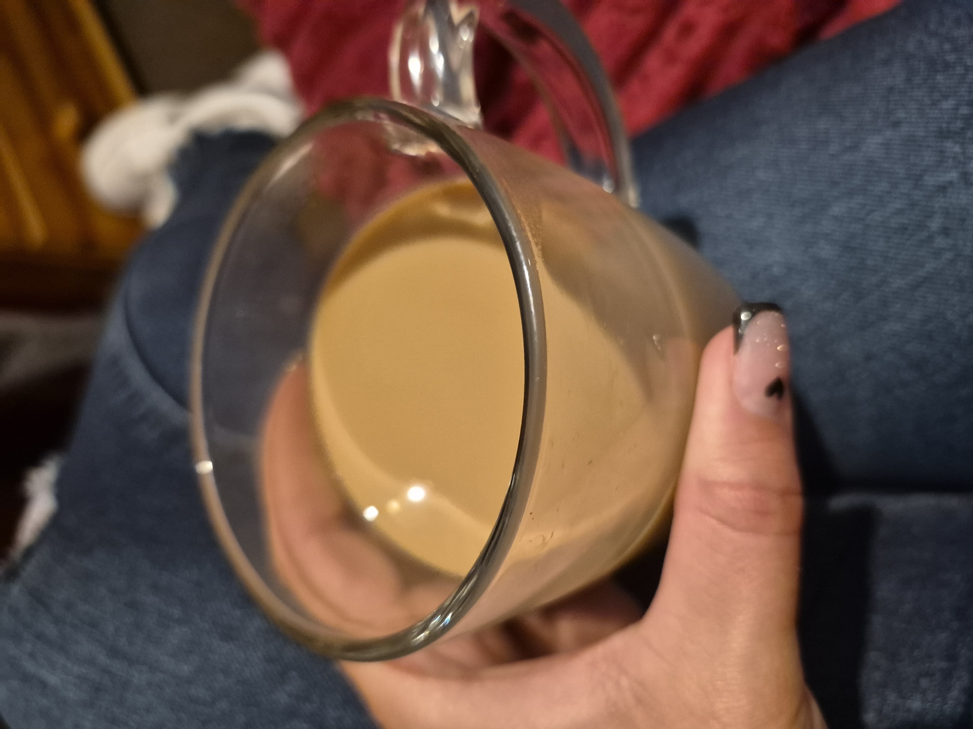 A close-up, eye-level shot shows a hand holding a clear glass mug filled with a light brown beverage, likely coffee with milk, resting on a person's lap, which is covered by dark blue denim jeans. The mug's handle is visible on the right side. In the background, a textured red fabric is partially visible. The scene does not provide any indicators of a specific location within Sandwell, United Kingdom.