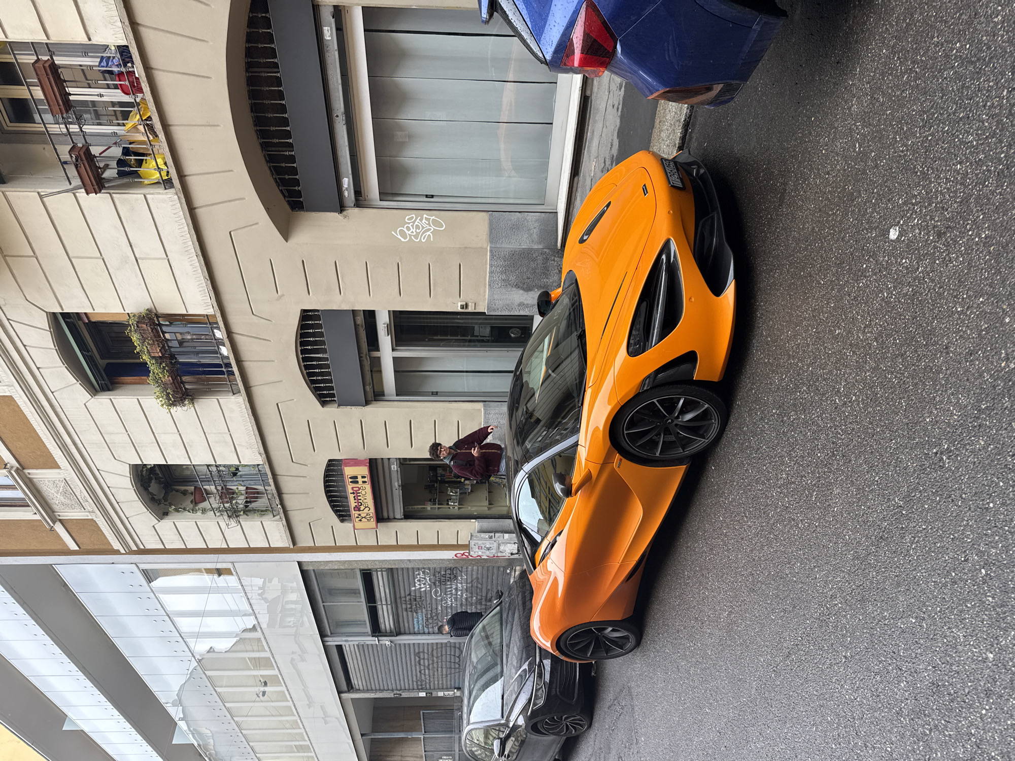A high-angle street-level view shows an orange McLaren sports car parked on the right side of a wet asphalt street in Milan, Italy. The front of a blue car is visible in the upper-right corner, and a dark-colored car is parked behind the McLaren. The street is lined with buildings featuring various architectural styles, including glass-paneled modern designs and older facades with balconies and wrought-iron details. A man stands in front of a store, holding a phone. Graffiti is visible on the walls of the buildings. The overall scene captures an urban setting with a luxury vehicle. The license plate indicates it’s foreign and includes the sequence ‘MD-063-5B’.
