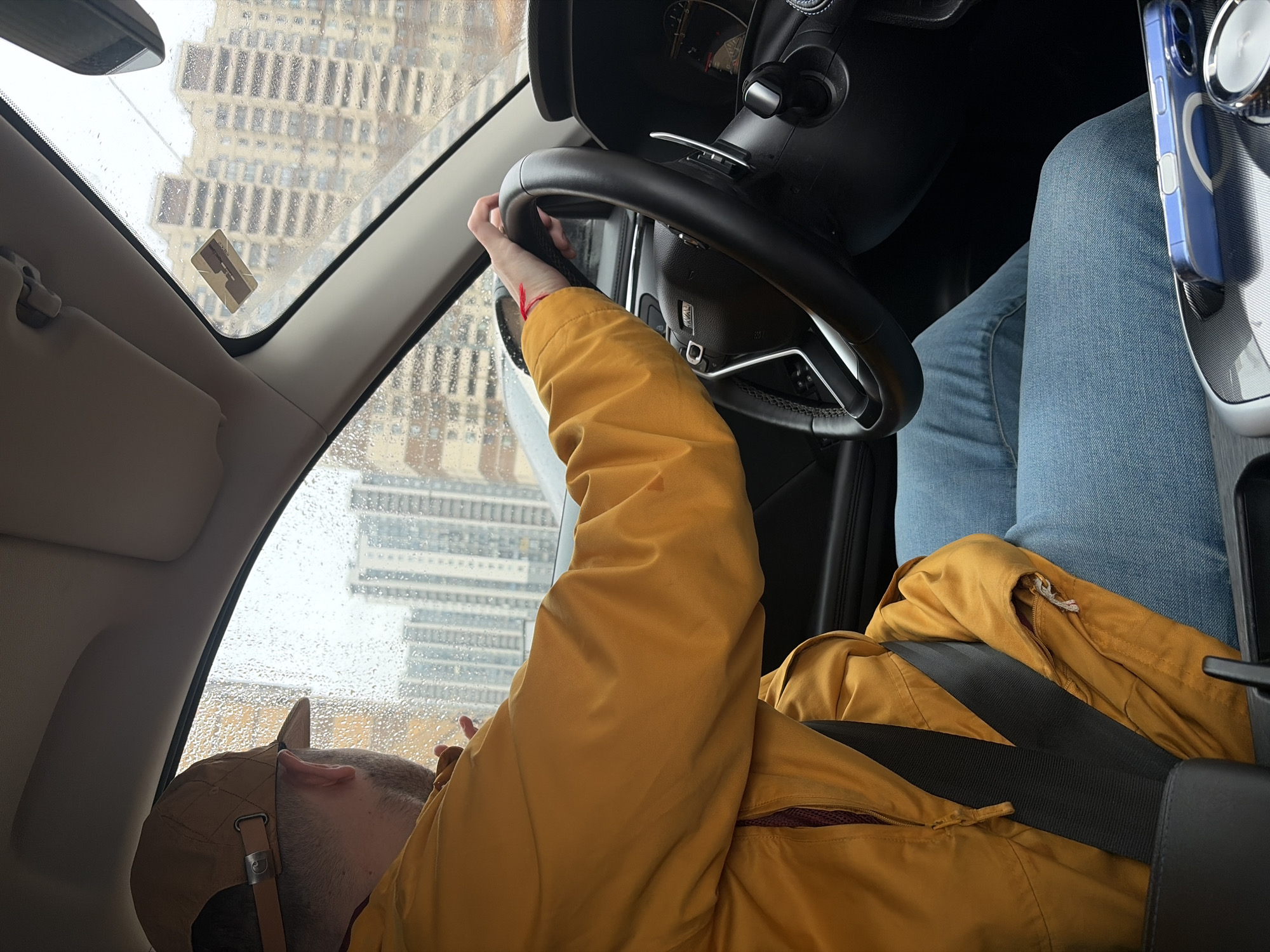 A man wearing a yellow jacket and a tan baseball cap lies reclined in the driver's seat of a car. He is wearing a seatbelt and has his left hand on the steering wheel. The car is in a city, with tall buildings visible through the rain-streaked windshield. A blue cell phone is mounted to the right of the steering wheel. Based on the architectural style of the building visible through the window, this image was taken in Saint Petersburg, Russia.