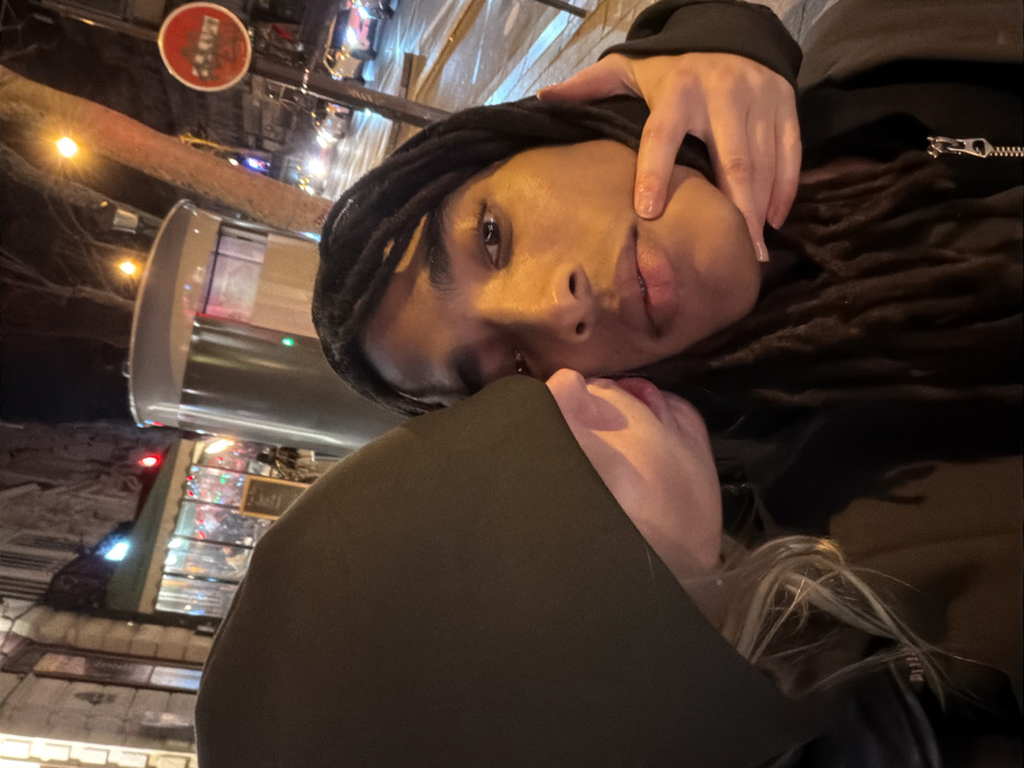 Two individuals pose closely together in a dimly lit urban environment. One, with dark skin, wears dreadlocks and a dark jacket with a zipper. A hand with painted nails touches their cheek. The other person, partially obscured by a dark hood, has fair skin and blonde hair. A sign is visible in the background, along with blurred lights and street scenes. The image appears to be taken with a tilted perspective.