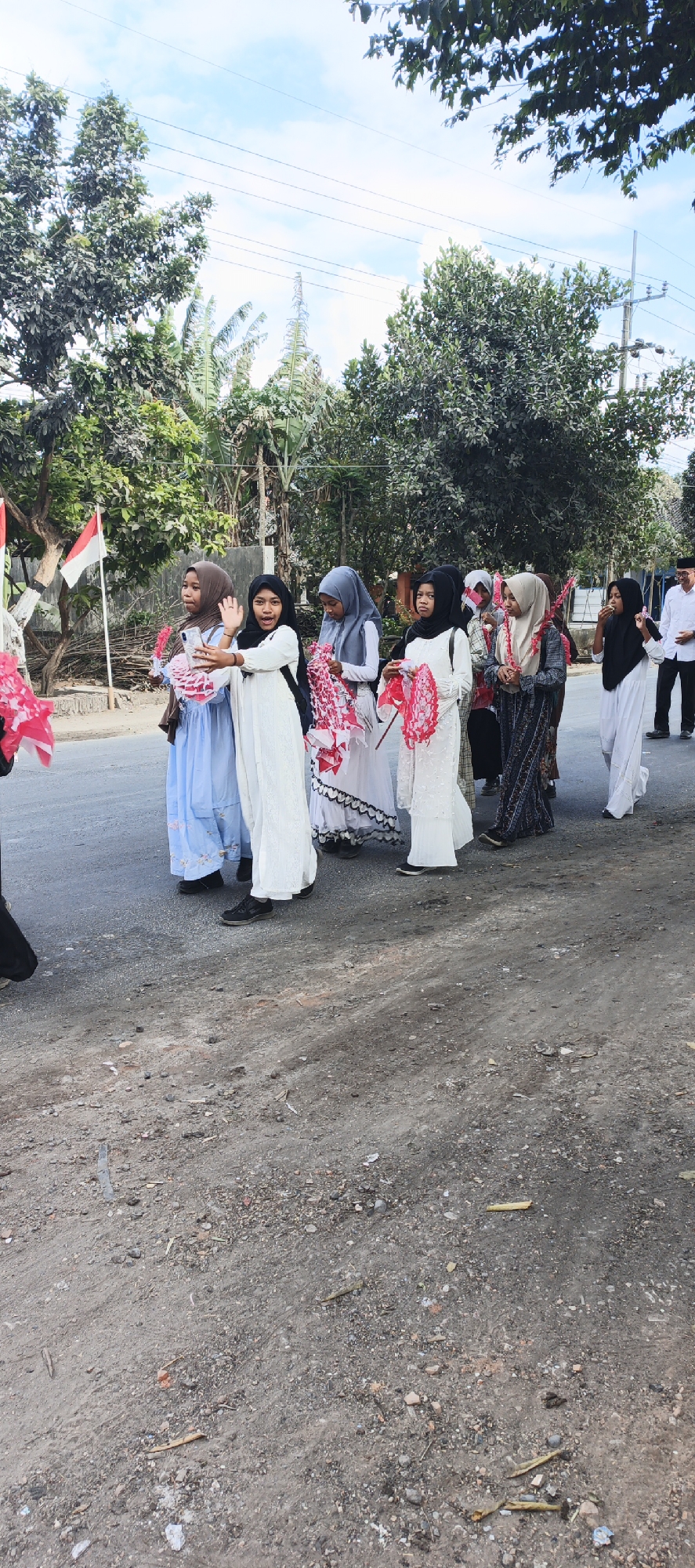 A group of young girls in hijabs and long dresses walk down a paved road. The girl in the front center is wearing a white dress and black hijab, holding a pink decorative object in her left hand and waving to the camera with her right. Other girls are wearing various colored hijabs and dresses, some also holding pink decorative objects. A man in a white shirt and black pants is visible in the background on the right, walking in the same direction as the girls. Two Indonesian flags are planted on the left side of the road. The environment appears to be a rural or suburban area with trees and utility poles visible.