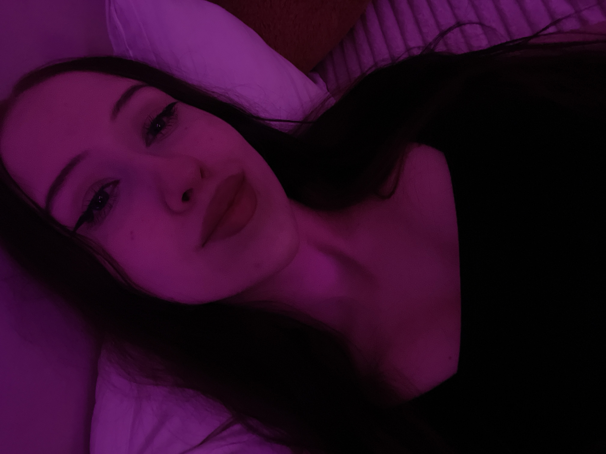 The image is a close-up selfie taken in Votkinsk, Russia. A young woman with long, dark hair is lying down, illuminated by pinkish-purple light. She is wearing a black top and has winged eyeliner. Her head is resting on a white pillow, and the background appears to be a textured surface, possibly a bedspread, also lit with the same pink light. The lighting creates a dramatic and intimate atmosphere.