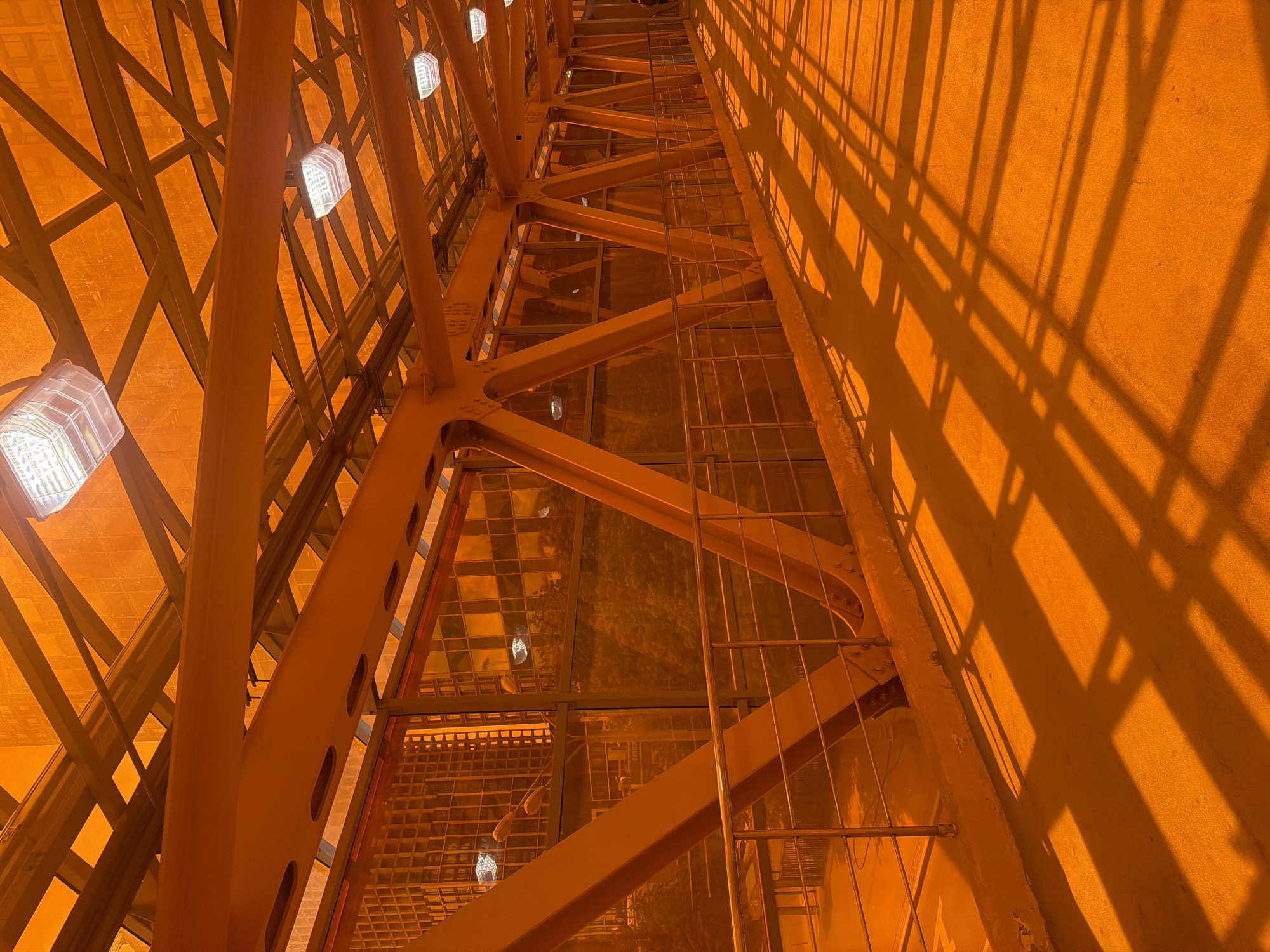 An interior view of an enclosed pedestrian bridge painted entirely in a vibrant orange hue. The bridge features a metal truss structure supporting an orange translucent roof and walls. Several rectangular light fixtures are mounted on the ceiling. The floor is also orange, and it is illuminated by long, geometric shadows cast by the bridge's structure from overhead lighting. A metal railing runs along the left side of the walkway. The background through the orange walls reveals blurred shapes of buildings and trees. A single person is visible walking in the distance on the right side of the bridge. The scene is bathed in an orange light, creating a warm and somewhat surreal atmosphere.